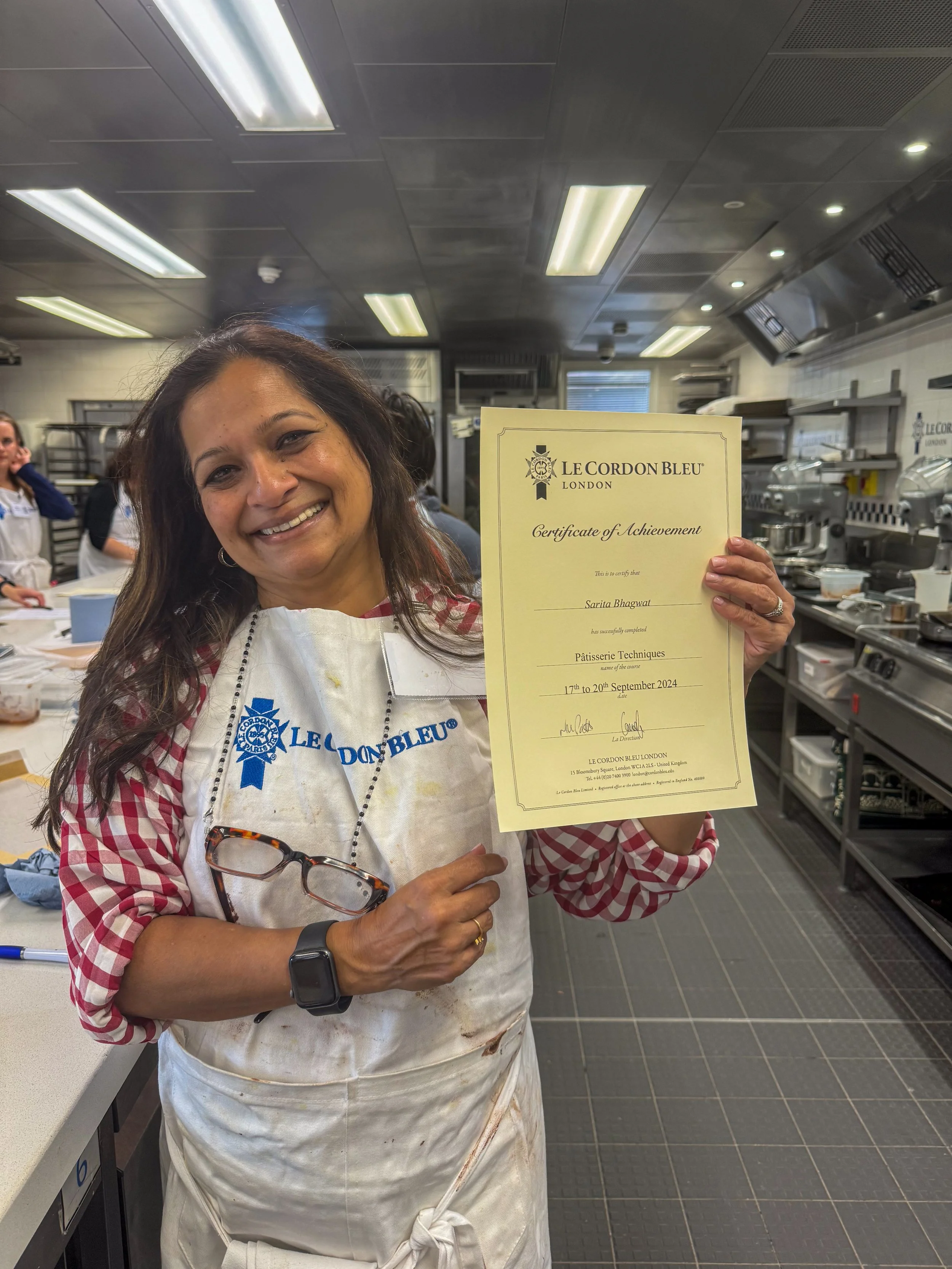 A woman wearing a white apron with blue text and a checkered shirt underneath, holding a certificate of achievement, smiling. She has glasses hanging on her shirt, a smartwatch, and a ring, and is in a professional kitchen setting with other people and kitchen equipment in the background.