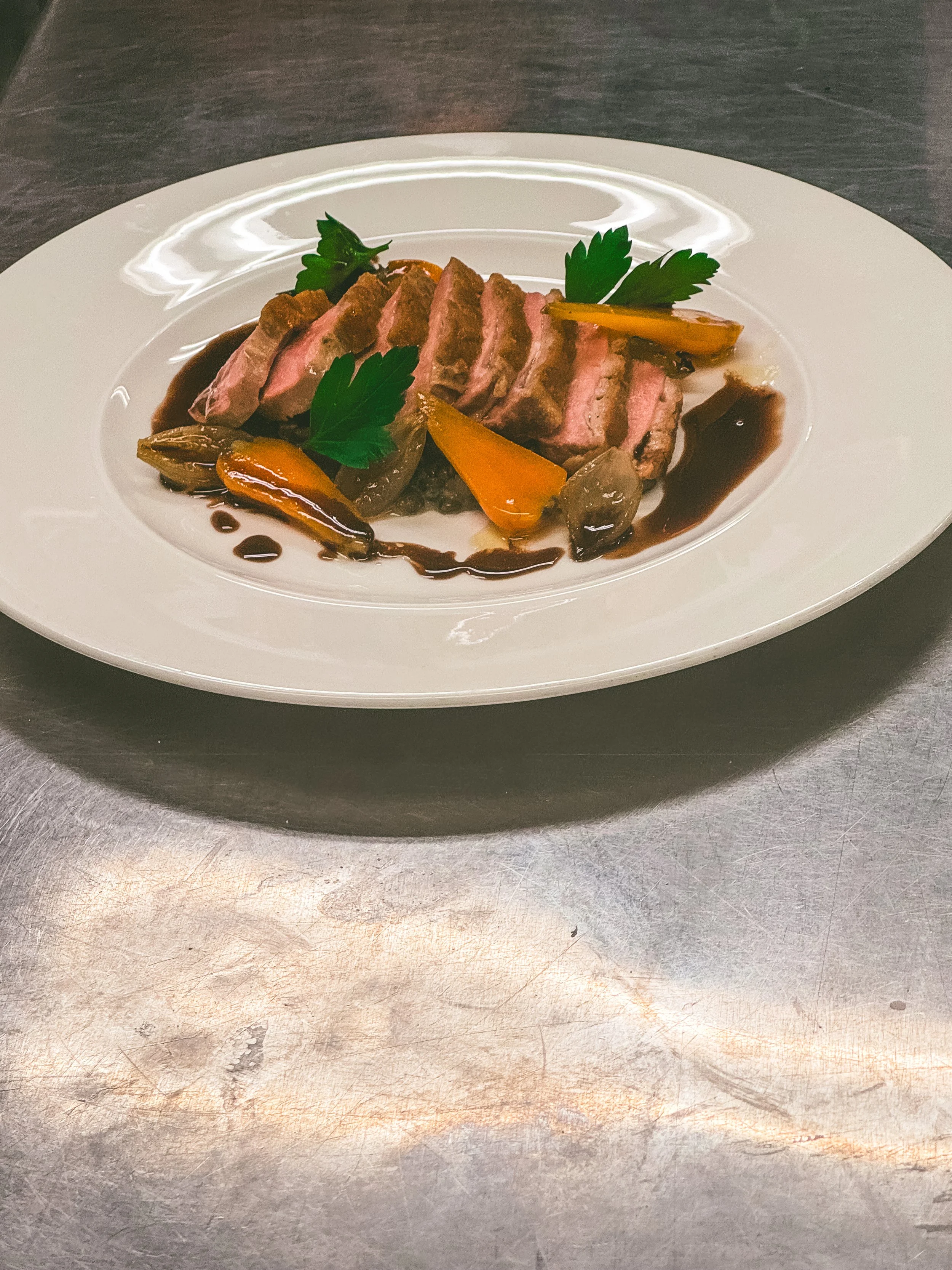 Gourmet plate with slices of cooked meat, roasted vegetables, fresh parsley, and a dark sauce drizzle, resting on a stainless steel surface.