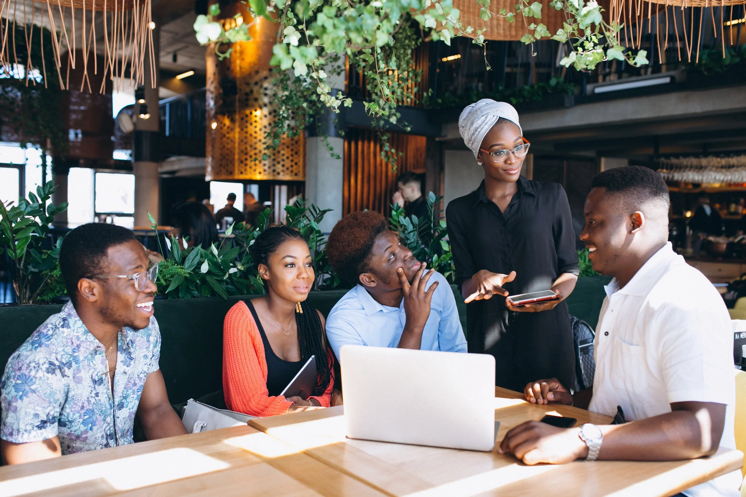 A group of five diverse people, four women and one man, are sitting around a table in a modern, well-lit restaurant or cafe, engaged in conversation. A server, a woman wearing glasses, a grey head wrap, and a black shirt, is standing beside them explaining or discussing something while holding a tablet.