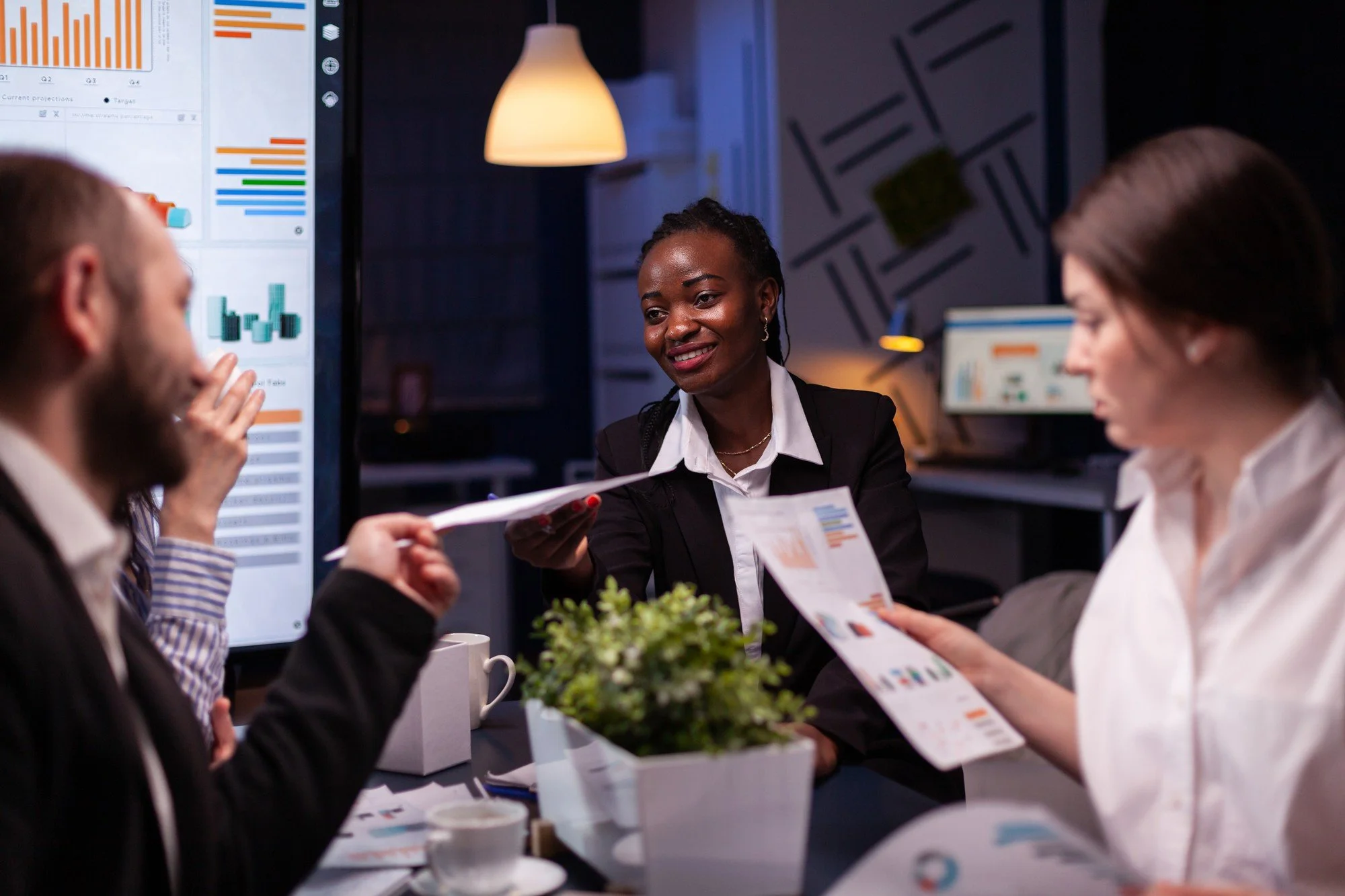 Business meeting in a conference room with four people, one woman smiling and holding a paper, others looking at charts on screens, with reports and coffee cups on the table.