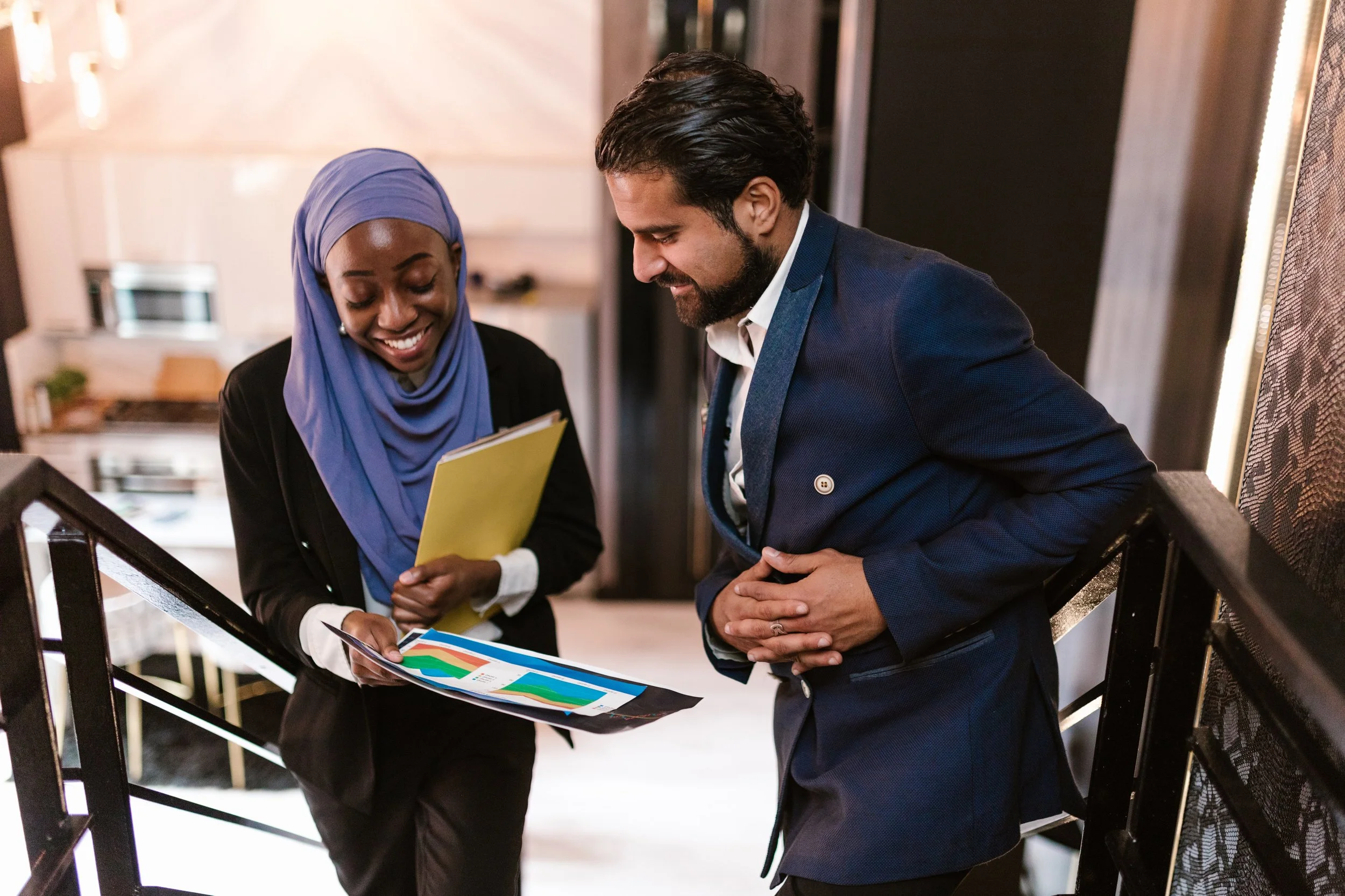 A woman wearing a blue headscarf and black blazer sharing a colorful printed document with a man in a navy blue suit inside a modern building.
