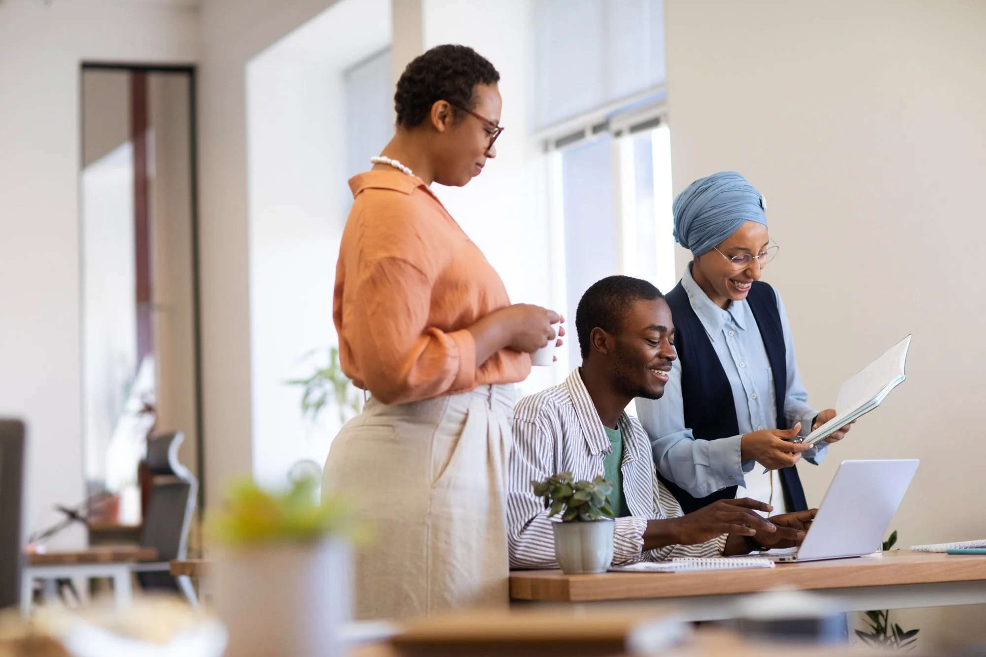 Three diverse coworkers, two women and one man, working together in an office. The woman on the left holds a coffee cup, the man is seated at a desk with a laptop, and the woman on the right holds a document, all smiling and engaged in collaboration.