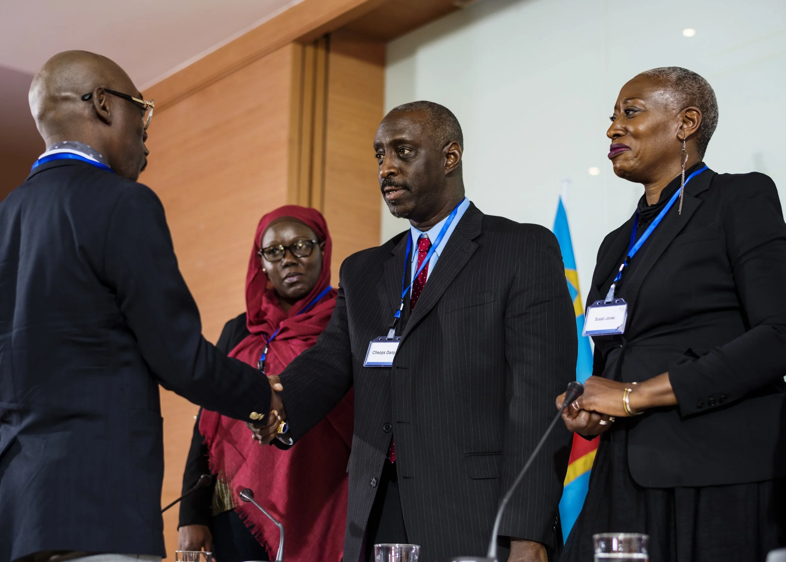 Two men and two women at a formal event, one woman wearing a red headscarf and glasses, the other woman with short gray hair and earrings, shaking hands, with name tags around their necks.