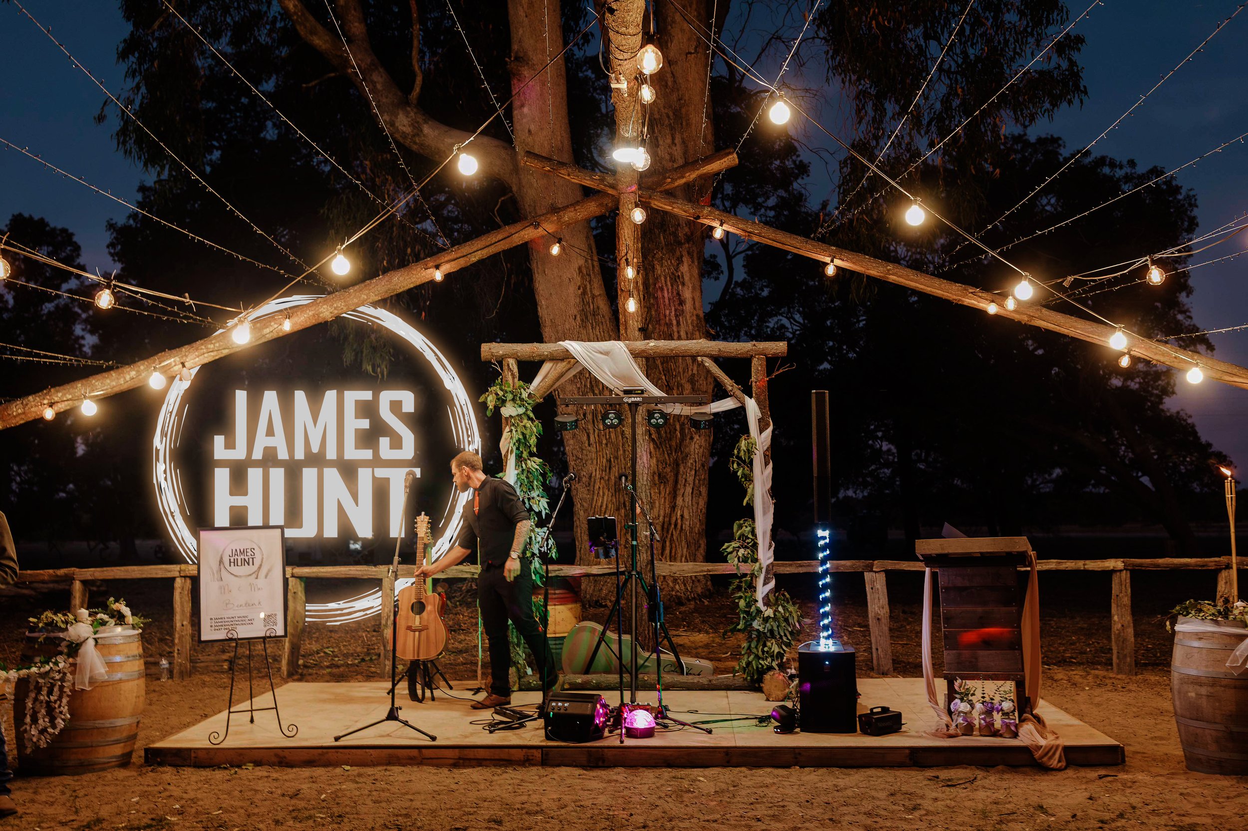 Outdoor wedding or event stage with a musician setting up under large trees decorated with string lights. A large illuminated sign reads 'James Hunt'. The stage is decorated with greenery, flowers, and a wooden frame, with microphones, guitars, speakers, and other musical equipment visible.