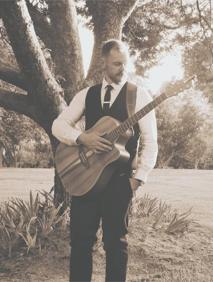 A man in formal attire, wearing a vest and tie, stands outdoors under a large tree, playing an acoustic guitar. The photo is in sepia tone.