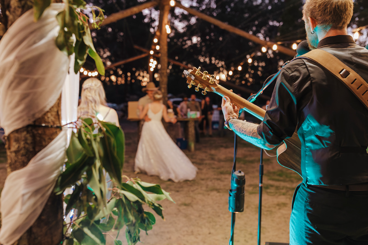 Hearts Afire Musician - James Hunt playing guitar at outdoor wedding reception during evening with guests and married couple in background. Located in Busselton