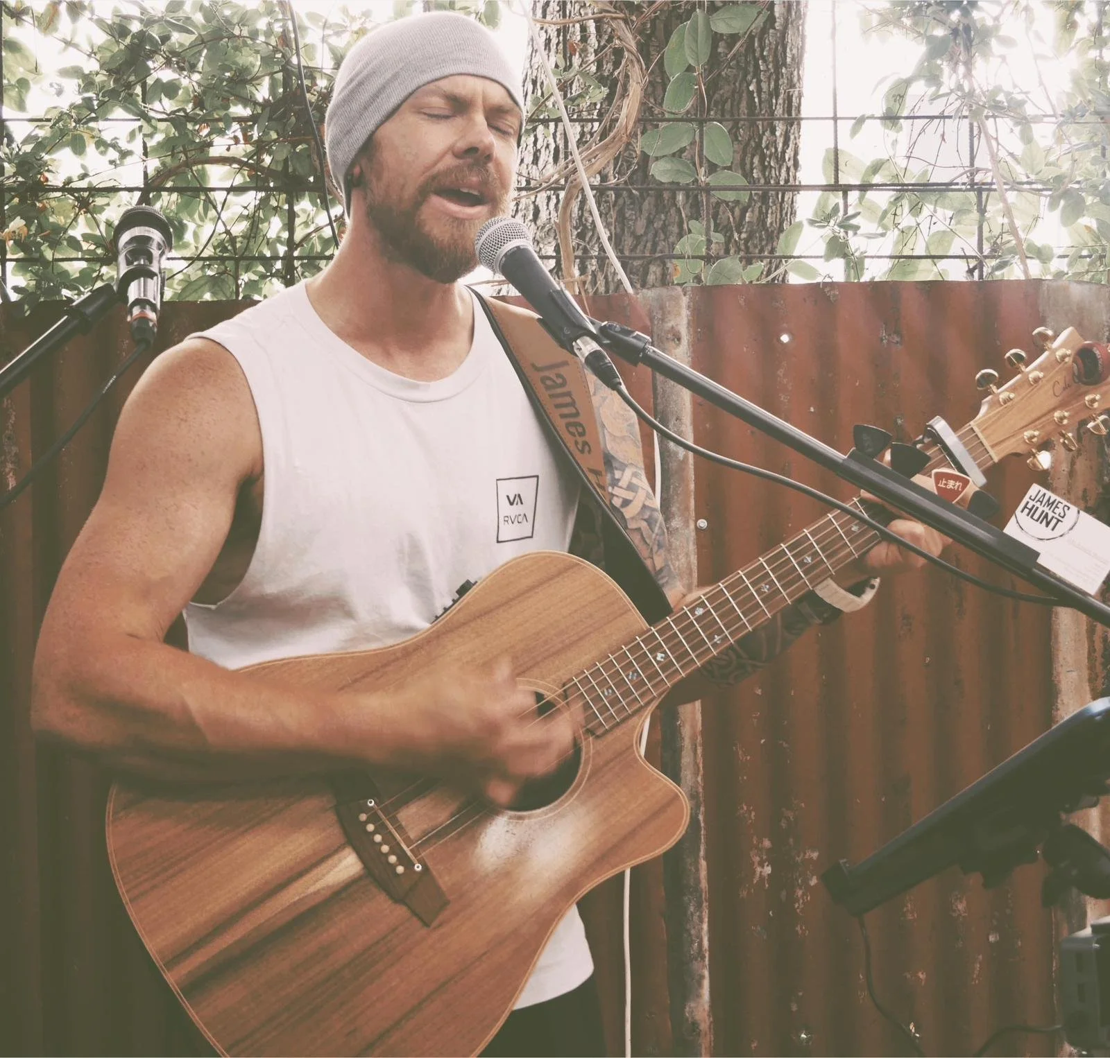 James Hunt from Hearts Afire is singing into a microphone while playing an acoustic guitar. The background shows a metal fence, a large tree trunk, and leafy foliage.