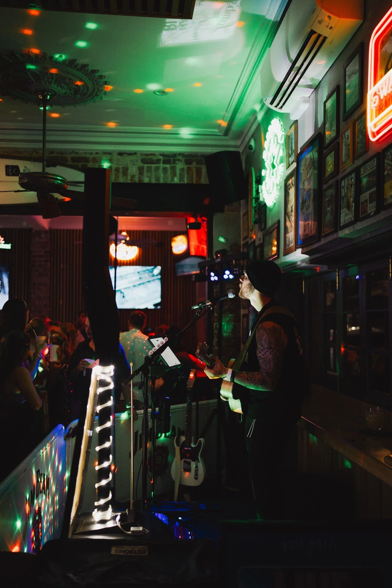 James Hunt from Hearts Afire playing guitar and singing into a microphone in a dimly lit bar or pub, with neon signs and framed pictures on the wall behind him, and a crowd of people watching.