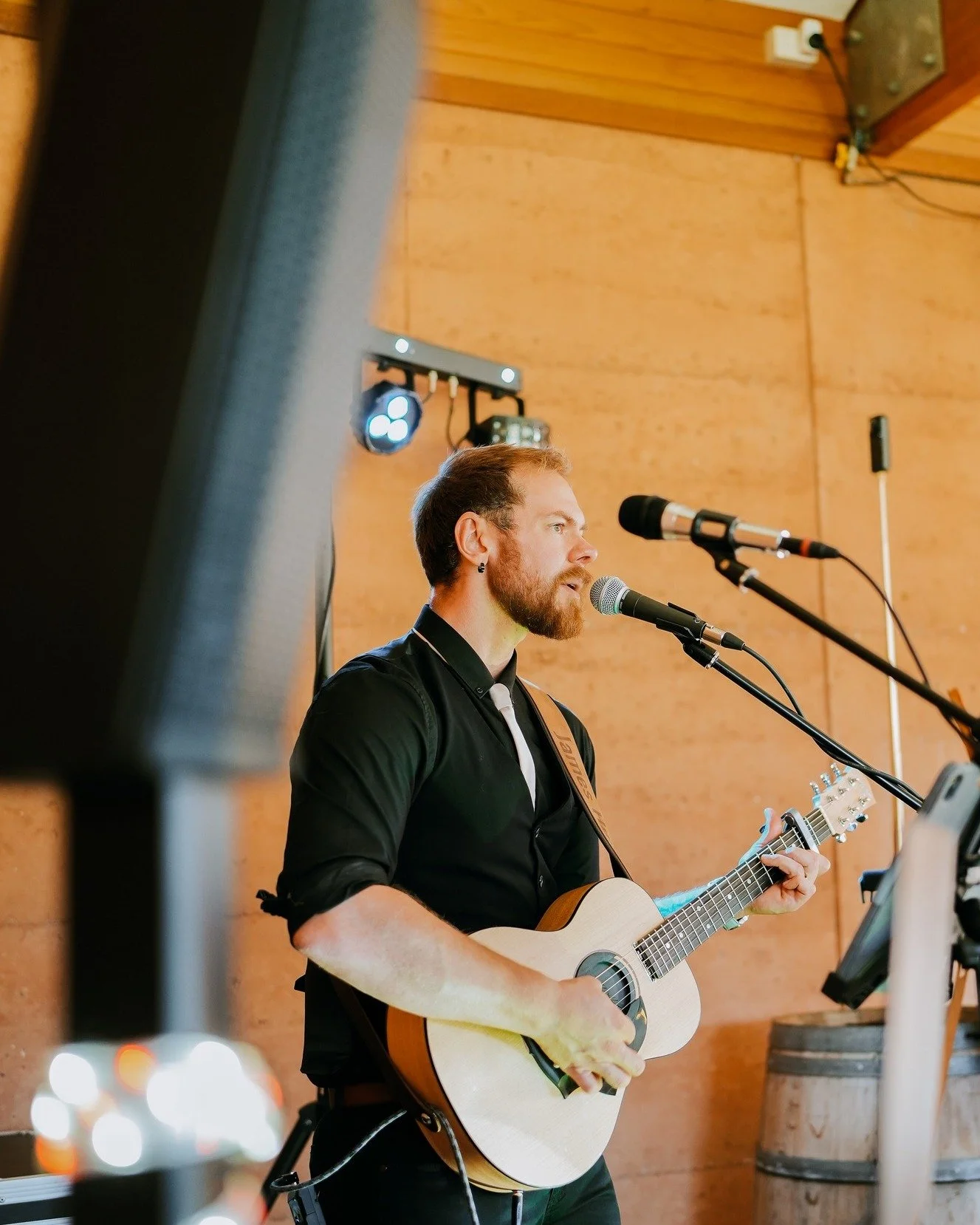 James from Hearts Afire is playing an acoustic guitar and singing into a microphone during a performance. The background consists of wooden panel walls, with sound equipment and lights visible.