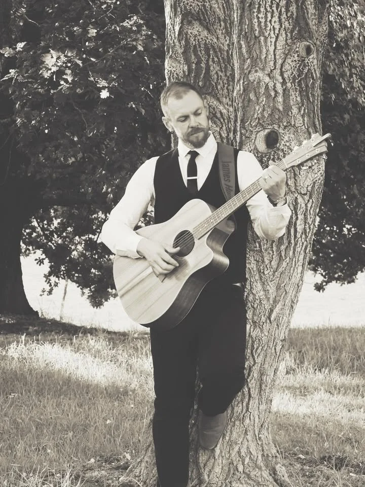 A man with a beard and mustache playing an acoustic guitar outdoors, standing beside a large tree in a grassy area with trees in the background.