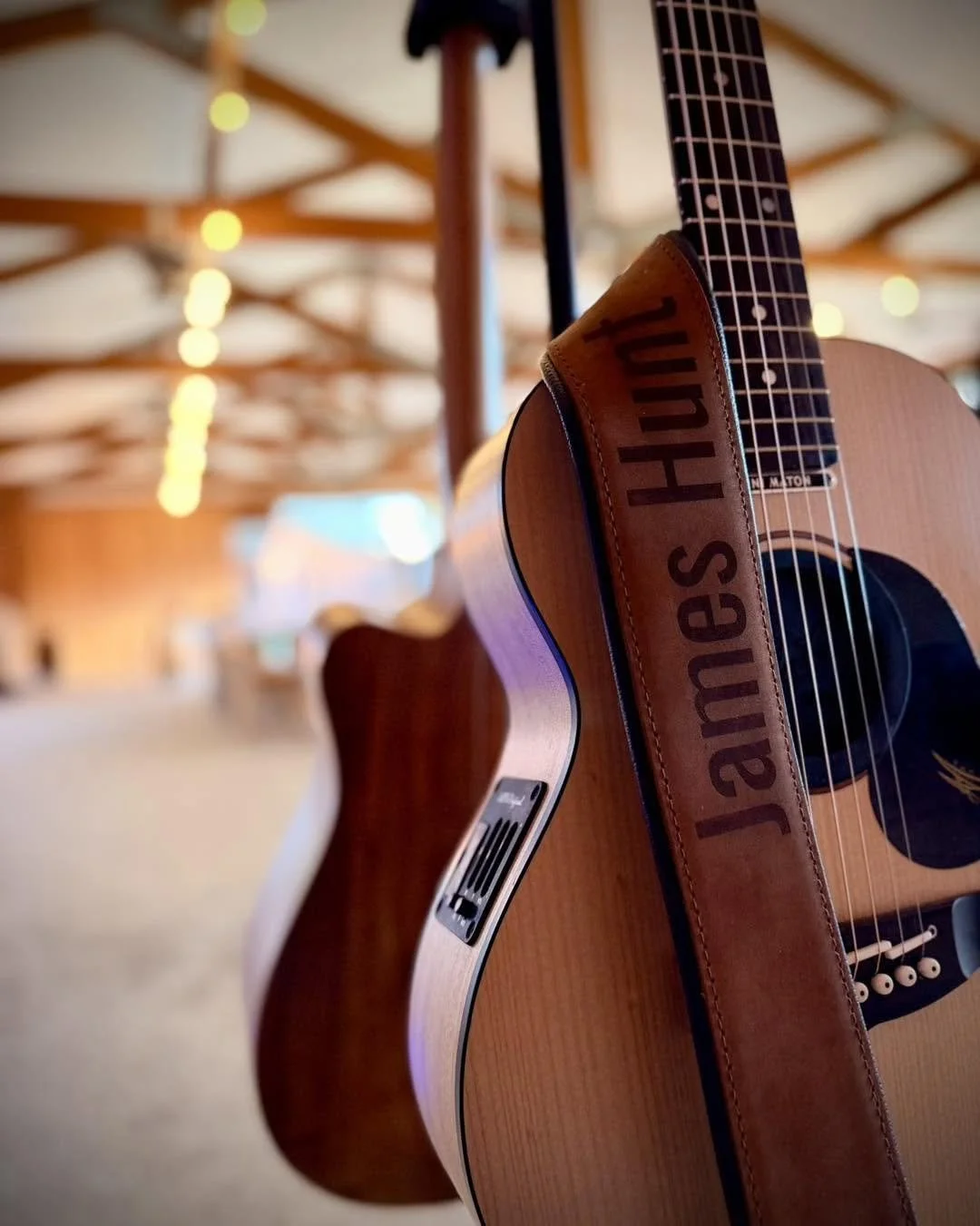 An acoustic guitar with a strap that says 'James H' hangs in a cozy, warmly lit space with wooden beams and string lights.