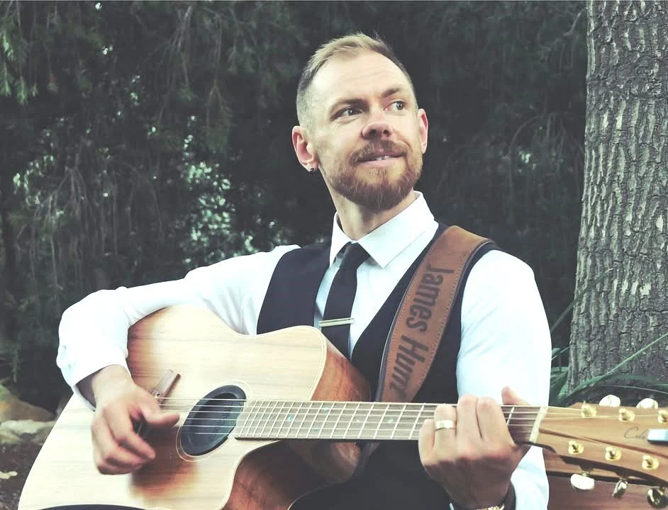 James Hunt, a musician, MC & DJ from Hearts Afire, a man with a beard and a short haircut playing an acoustic guitar outdoors, dressed in a white shirt, black vest, and black tie, standing next to a tree. Located south of Perth