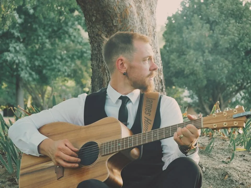 James Hunt, a musician, MC & DJ from Hearts Afire with short blond hair and a beard plays an acoustic guitar while sitting outdoors near a tree, dressed in a white shirt, black vest, and black tie. Located south of Perth