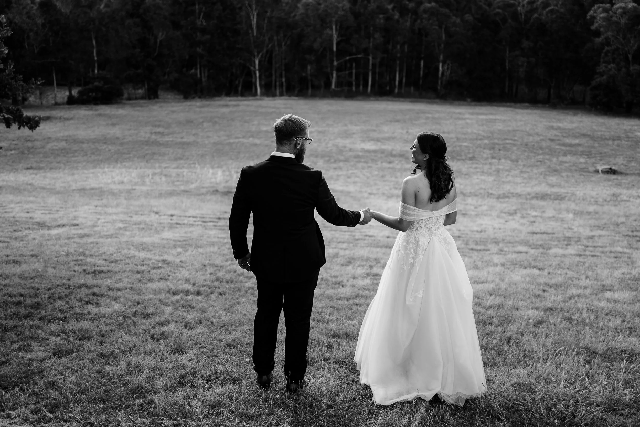 Black and white photo of a bride and groom standing on a grassy field, holding hands, with trees in the background.