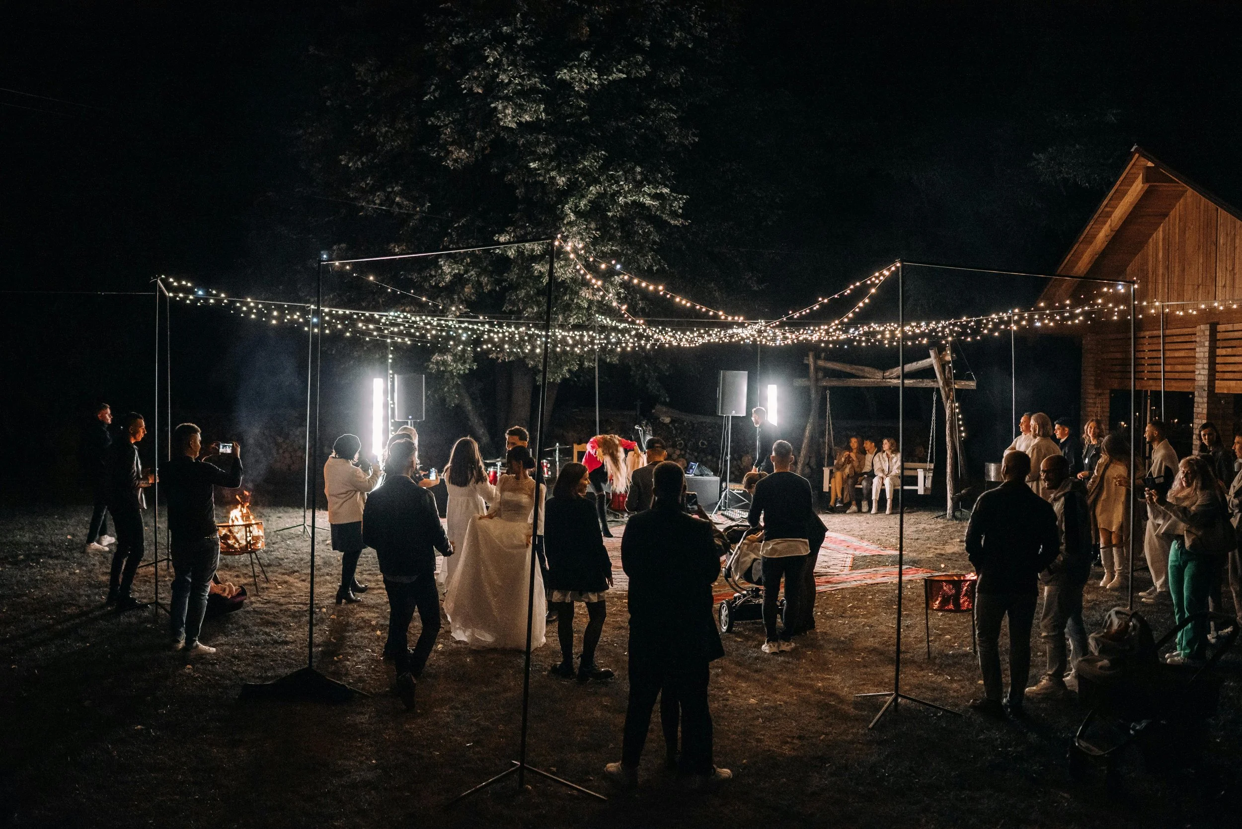 Nighttime outdoor celebration with string lights overhead and people dancing on a dirt ground, with a wooden building and some seated guests in the background.