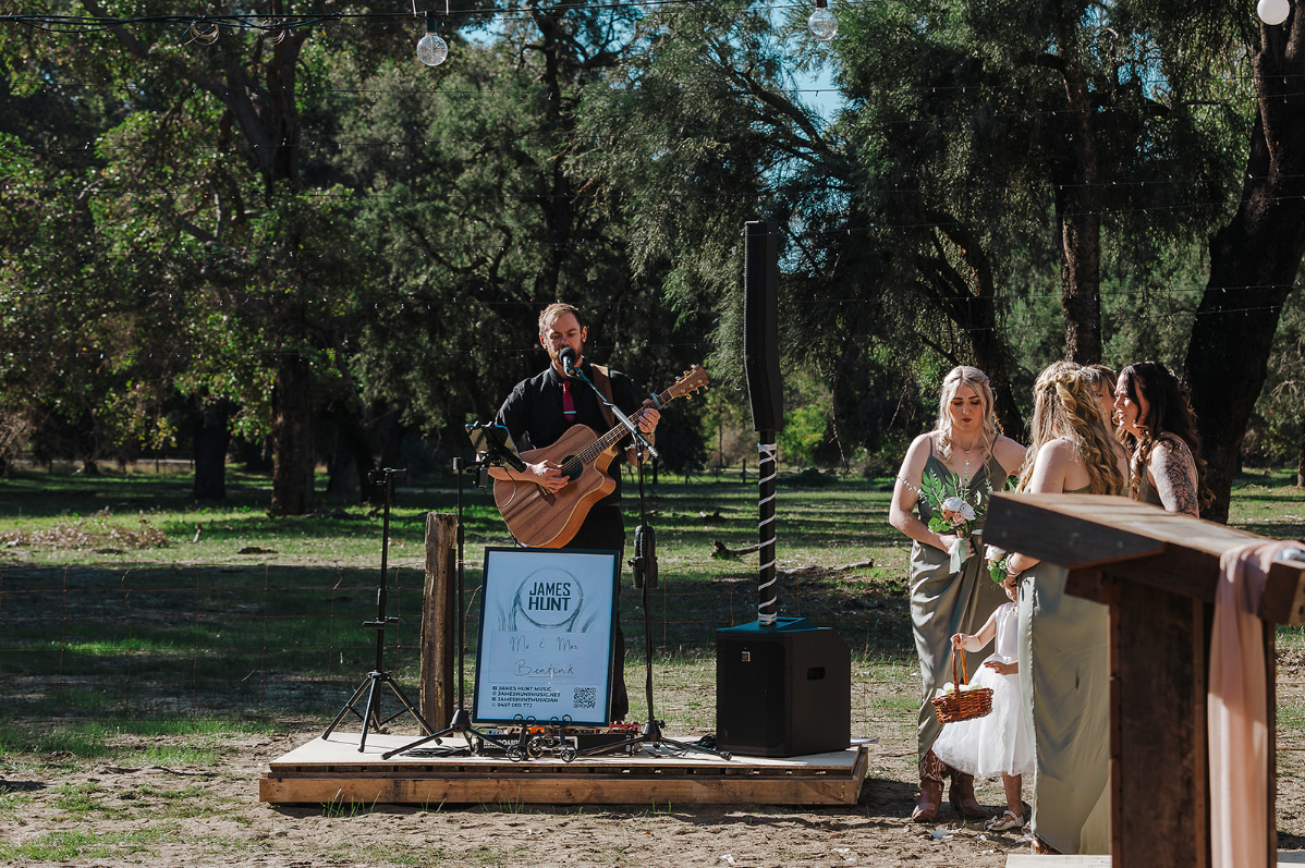 Hearts Afire Musician - James Hunt playing guitar and singing at outdoor wedding ceremony, with three women and a girl in wedding dress nearby under trees on sunny day. Located in Busselton