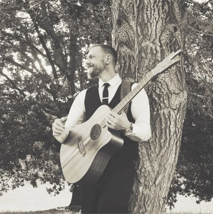 James Hunt, a musician, MC & DJ from Hearts Afire in formal attire playing an acoustic guitar outdoors, leaning against a tree with a smile. Located south of Perth