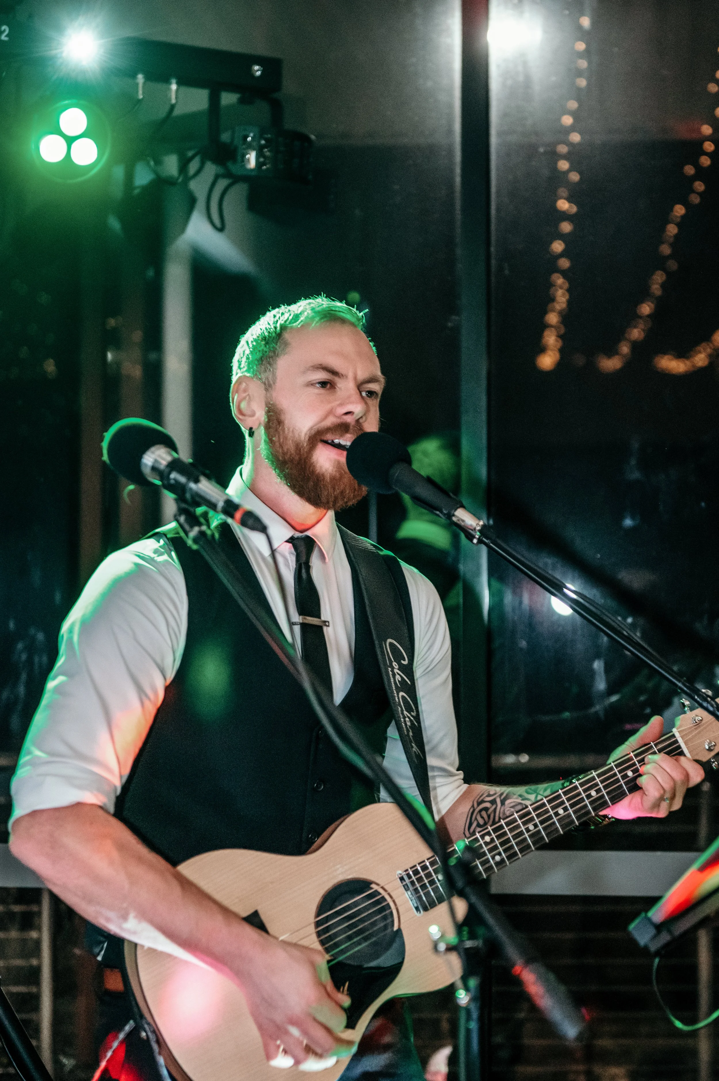 James Hunt from Hearts Afire is singing and playing an acoustic guitar at a live music performance, with microphones in front of him, a green stage light, and string lights in the background.