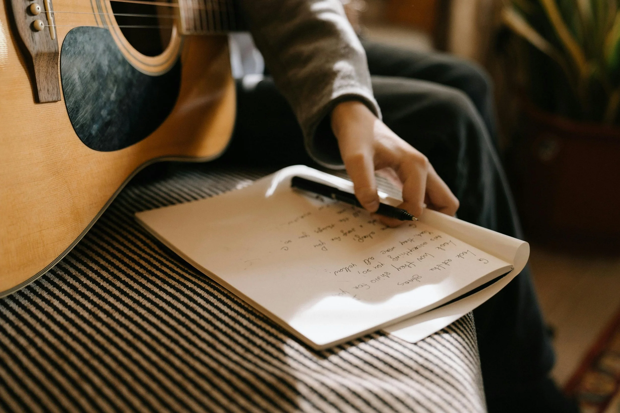 A person sitting with a guitar, writing in a notebook with a black pen.