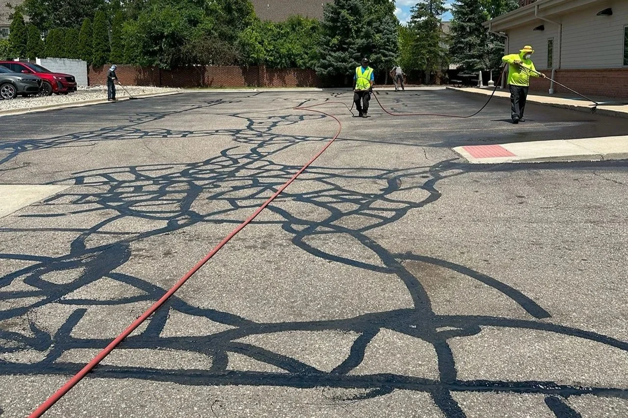 Workers applying fresh blacktop to a cracked parking lot surface, with power sprayers and workers in yellow safety vests.
