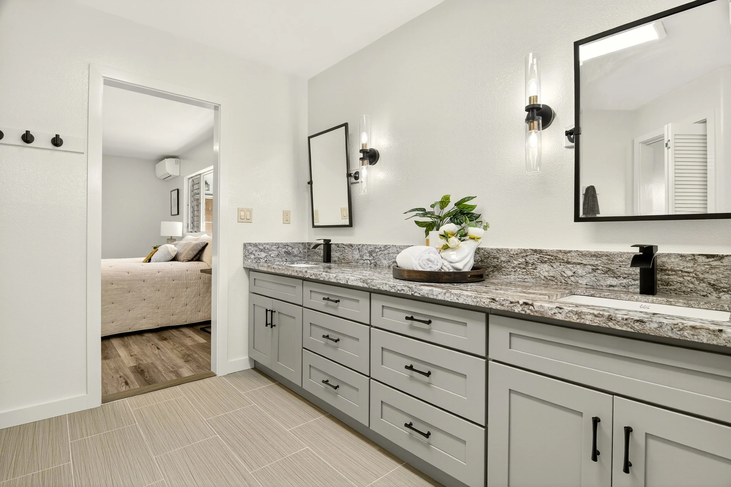 Modern bathroom with double vanity, granite countertops, and black fixtures, leading to a bedroom with a bed, lamps, and window