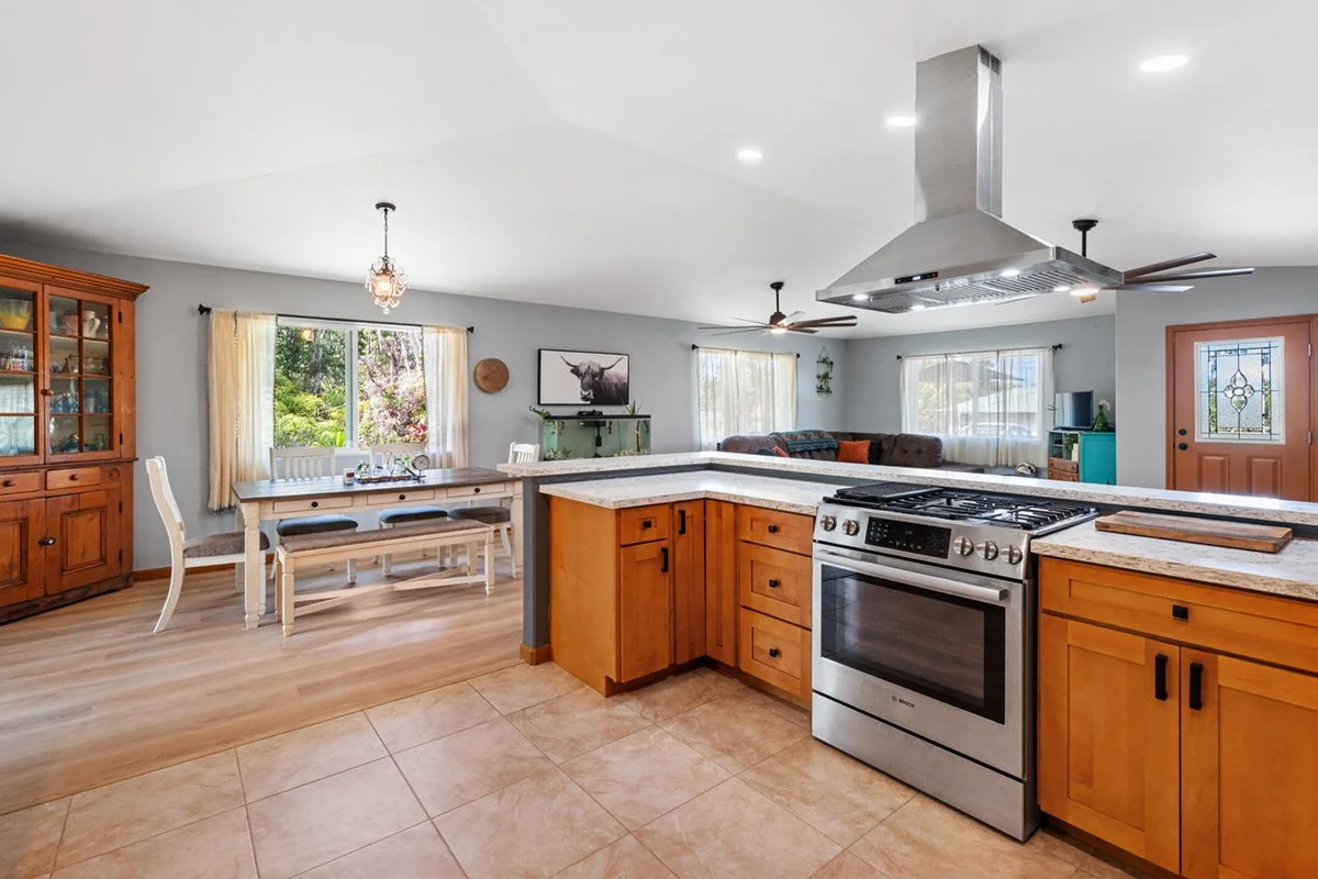 Open-concept kitchen and dining area with wooden cabinetry, a granite countertop, stainless steel stove, and a living room with sofas and windows showing greenery outside.
