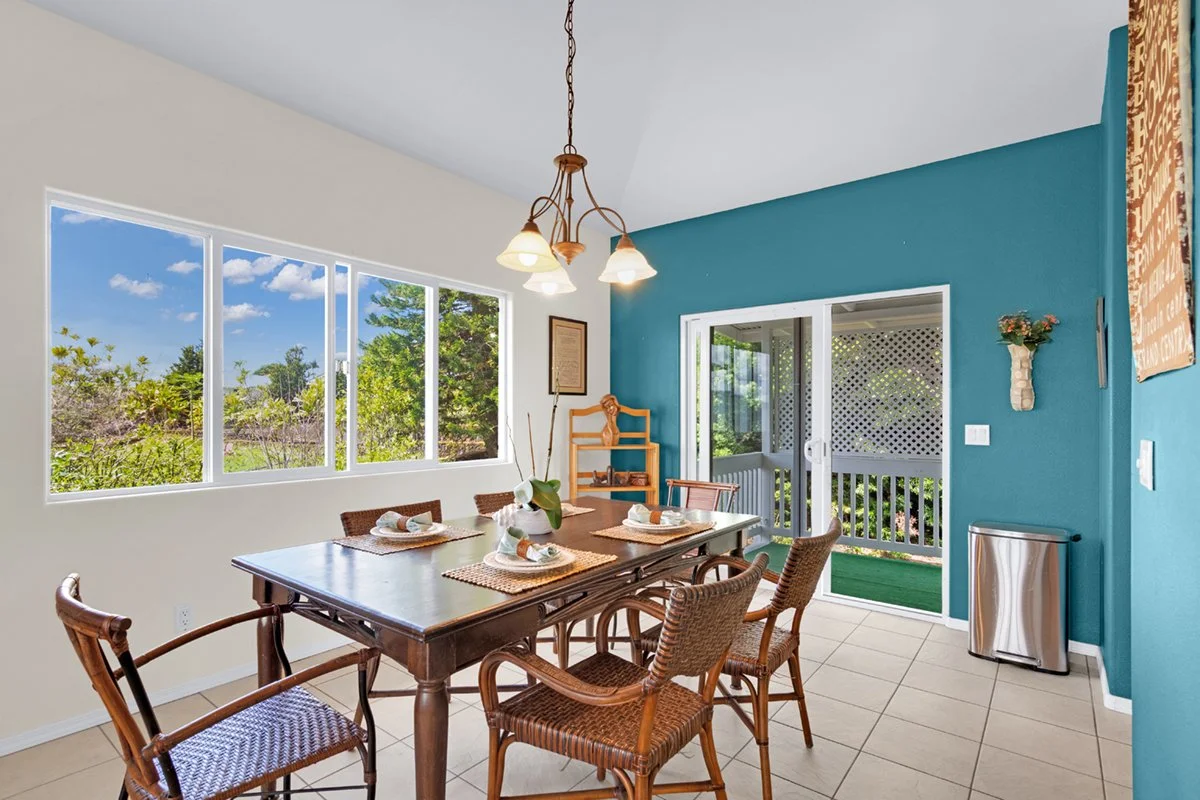 A dining room with a wooden table and six wicker chairs, set with plates, napkins, and paper towels. The room has a large window showing a blue sky and green trees, a blue accent wall, a sliding glass door to a porch, a hanging light fixture, a small shelf, and a trash can.