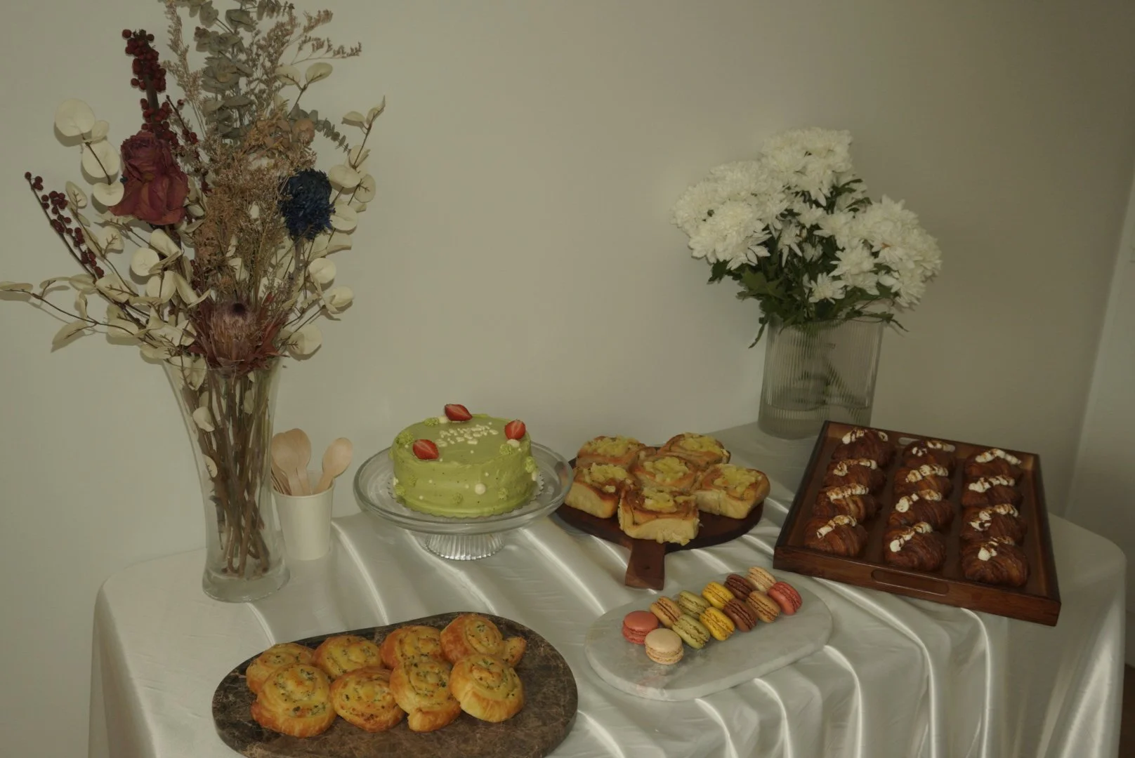 Table with various desserts, including a green cake, cookies, macarons, croissants, and slices of pie, decorated with two large flower arrangements in vases.