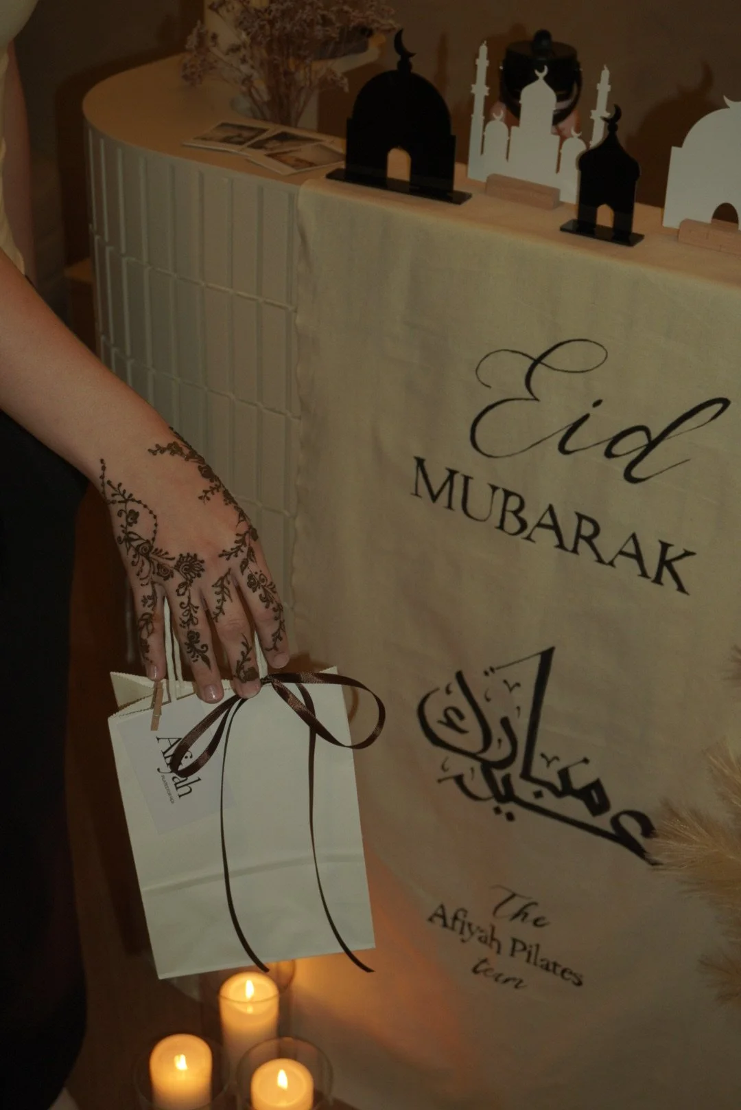 Person holding a gift bag with henna decorated hand, at an Eid celebration with a sign that reads 'Eid MUBARAK' and an Arabic greeting, Arabic calligraphy, candles, and henna decorations in the background.