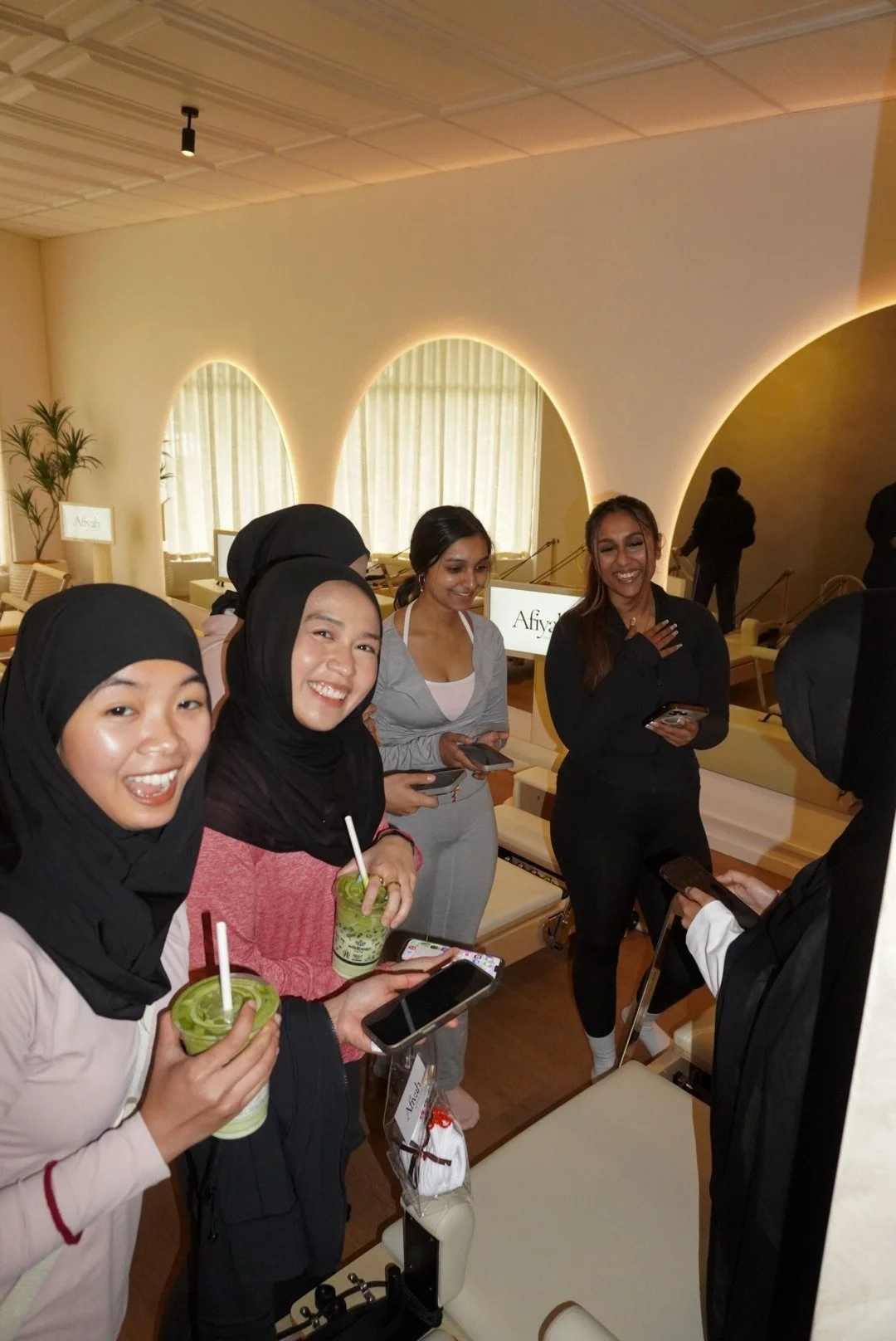 Group of diverse women smiling and holding drinks and phones in a modern, well-lit room with beige walls and large windows.