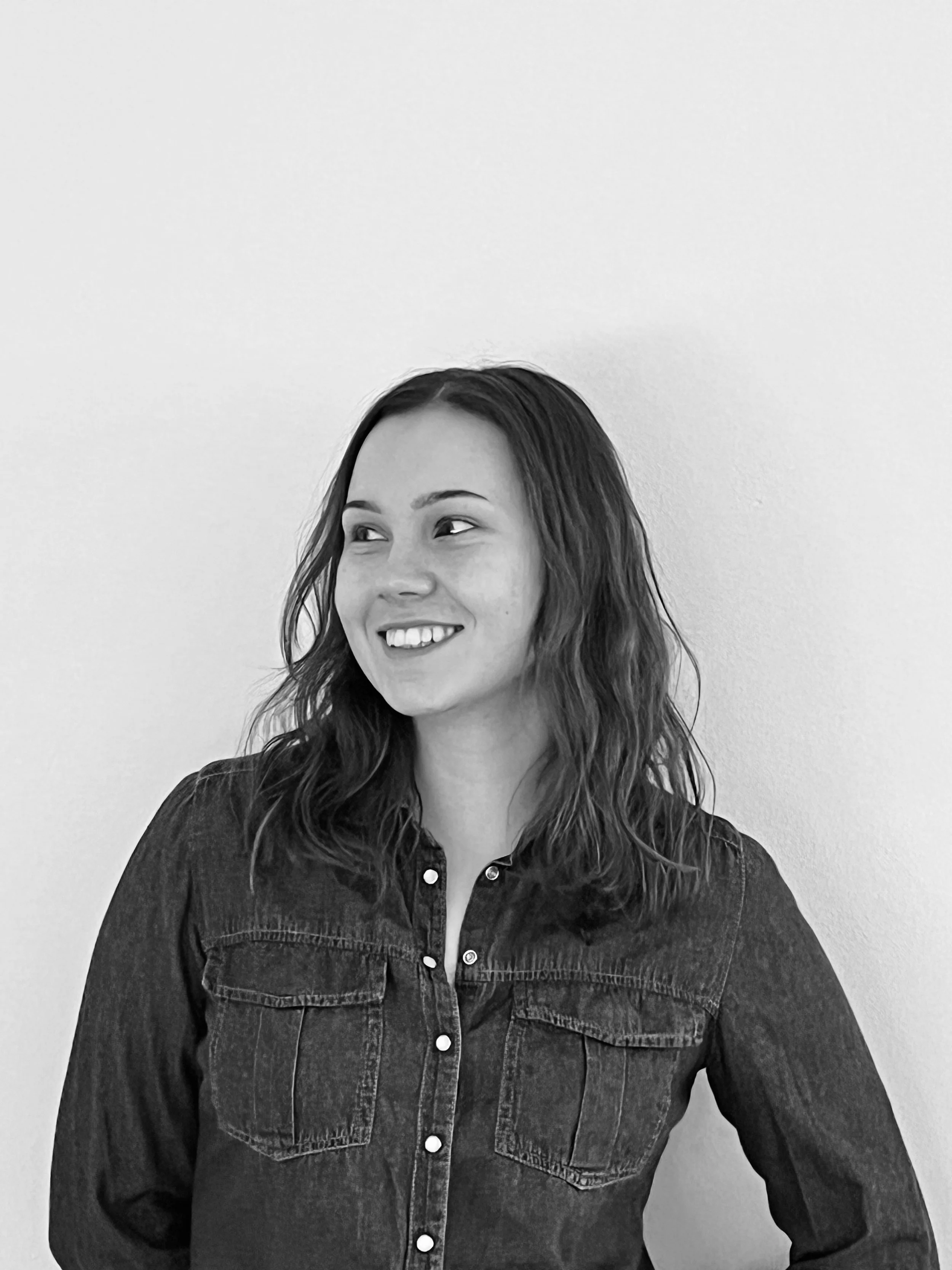 A smiling woman with long, wavy hair wearing a button-up denim shirt standing against a plain white wall.