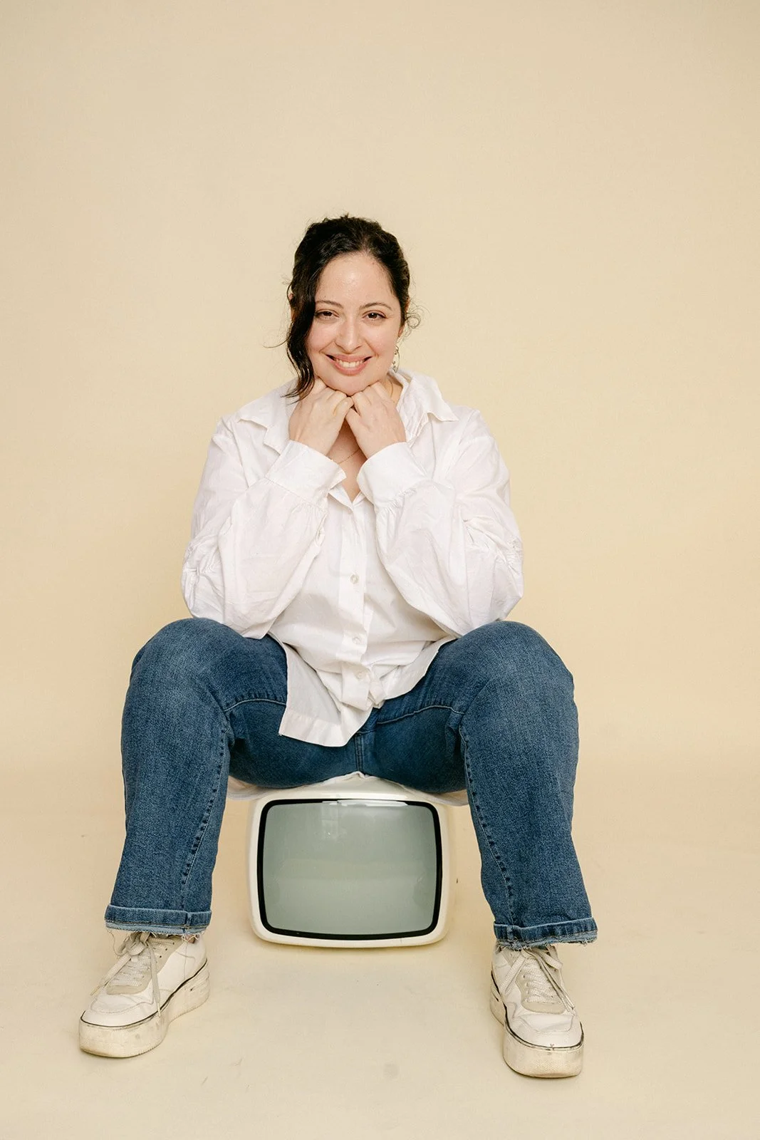 Femme souriante assise sur un tabouret en bois dans une pièce aux murs blancs avec des moulures, portant une robe beige en satin.