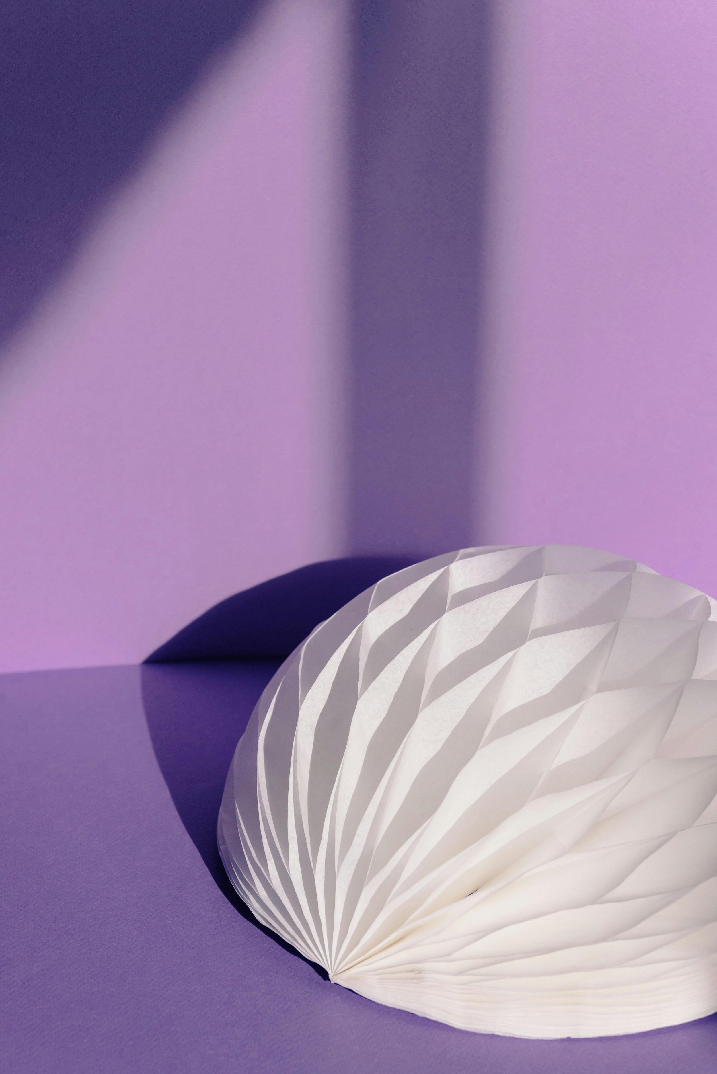 Une lampe en papier blanc avec un motif géométrique, posée sur une surface mauve, avec un fond violet et une ombre projetée en diagonale.