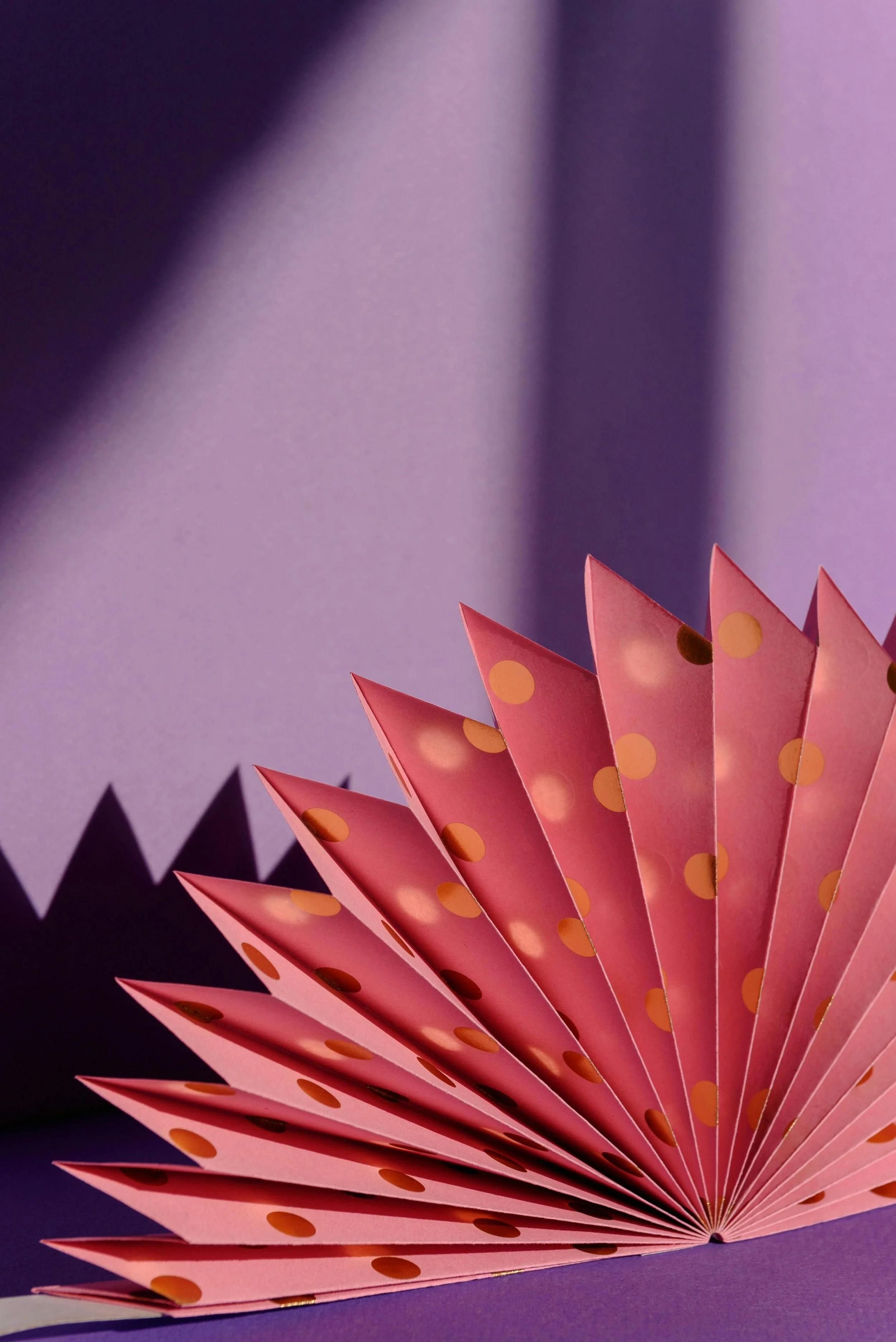 Fête de papier rose avec des points orange, pliée en forme d'éventail, sur un fond violet. Partiellement visible dans le coin inférieur gauche, un autre éventail violet.