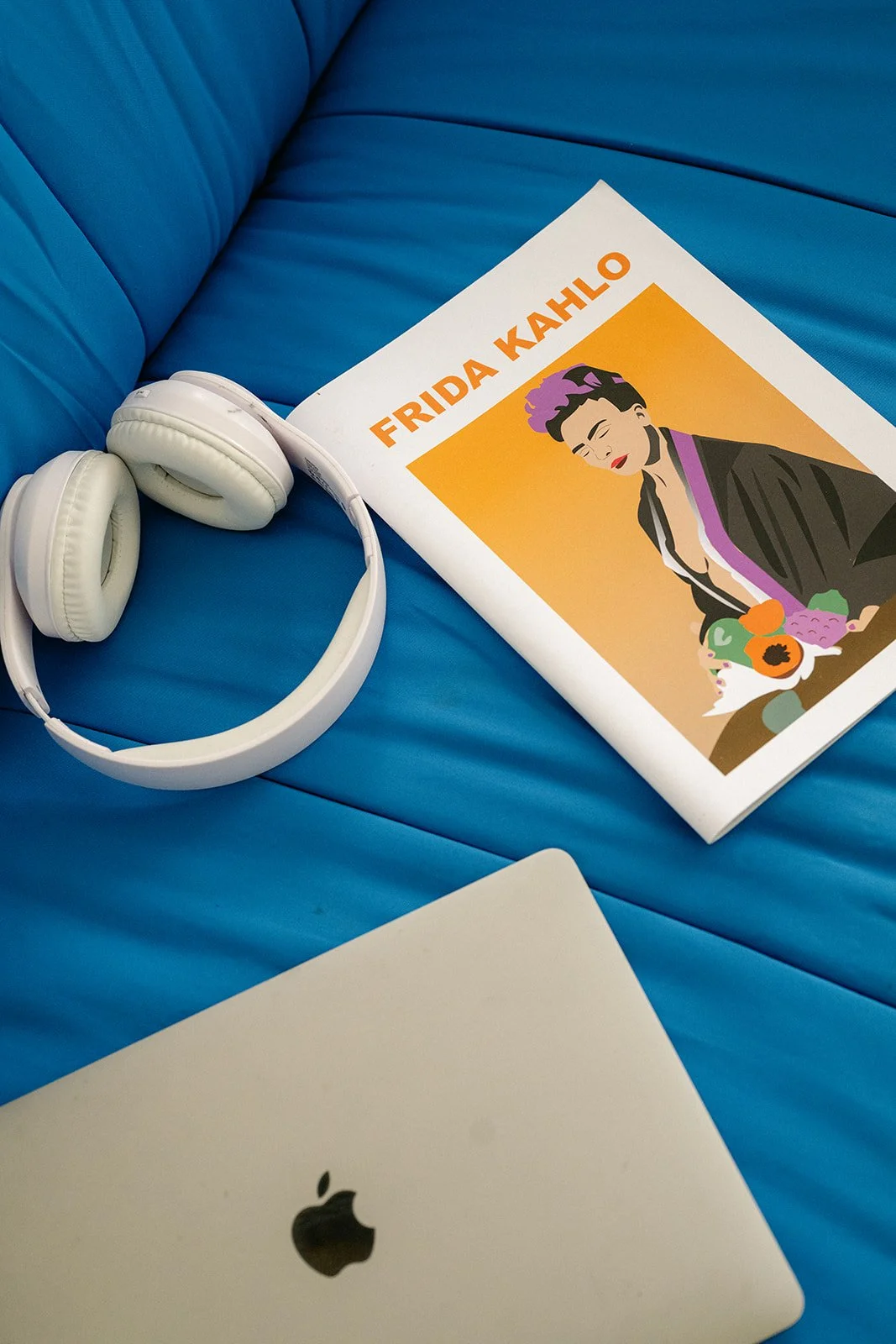 Une paire d'écouteurs blancs, un livre avec le titre 'FRIDA KAHLO' et une illustration de Frida Kahlo, et un ordinateur portable Apple, tous posés sur un canapé en cuir bleu.