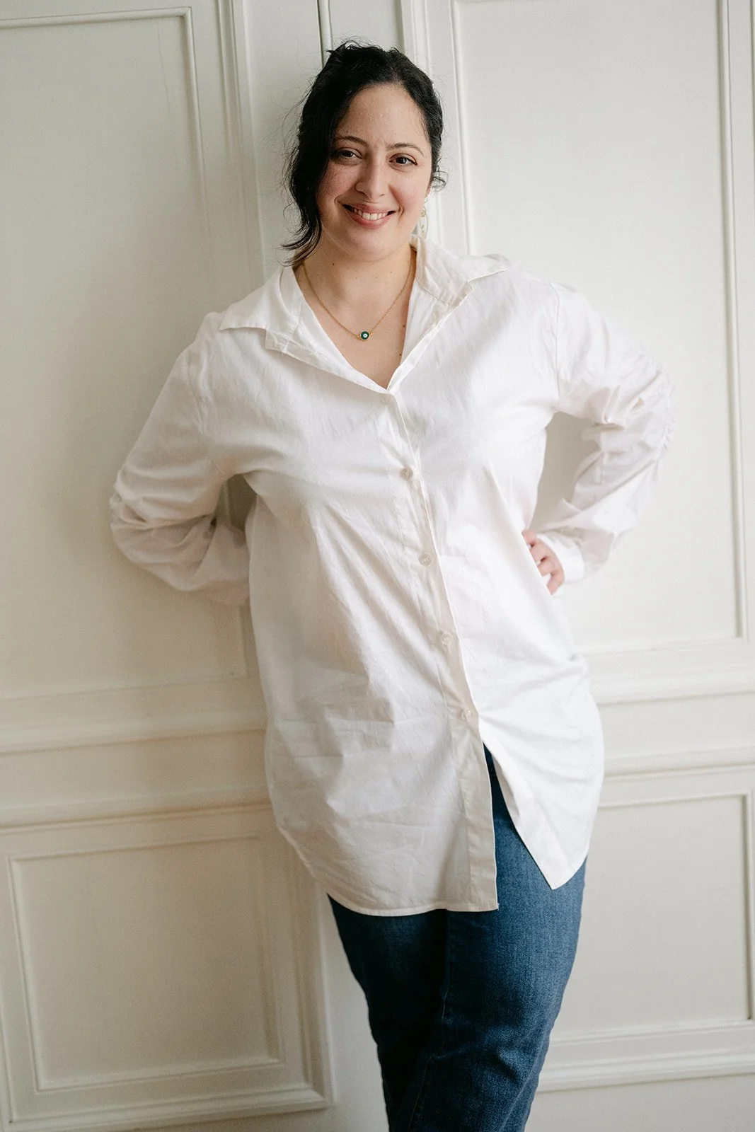 Femme souriante portant une chemise blanche et un jean, posant contre un mur blanc