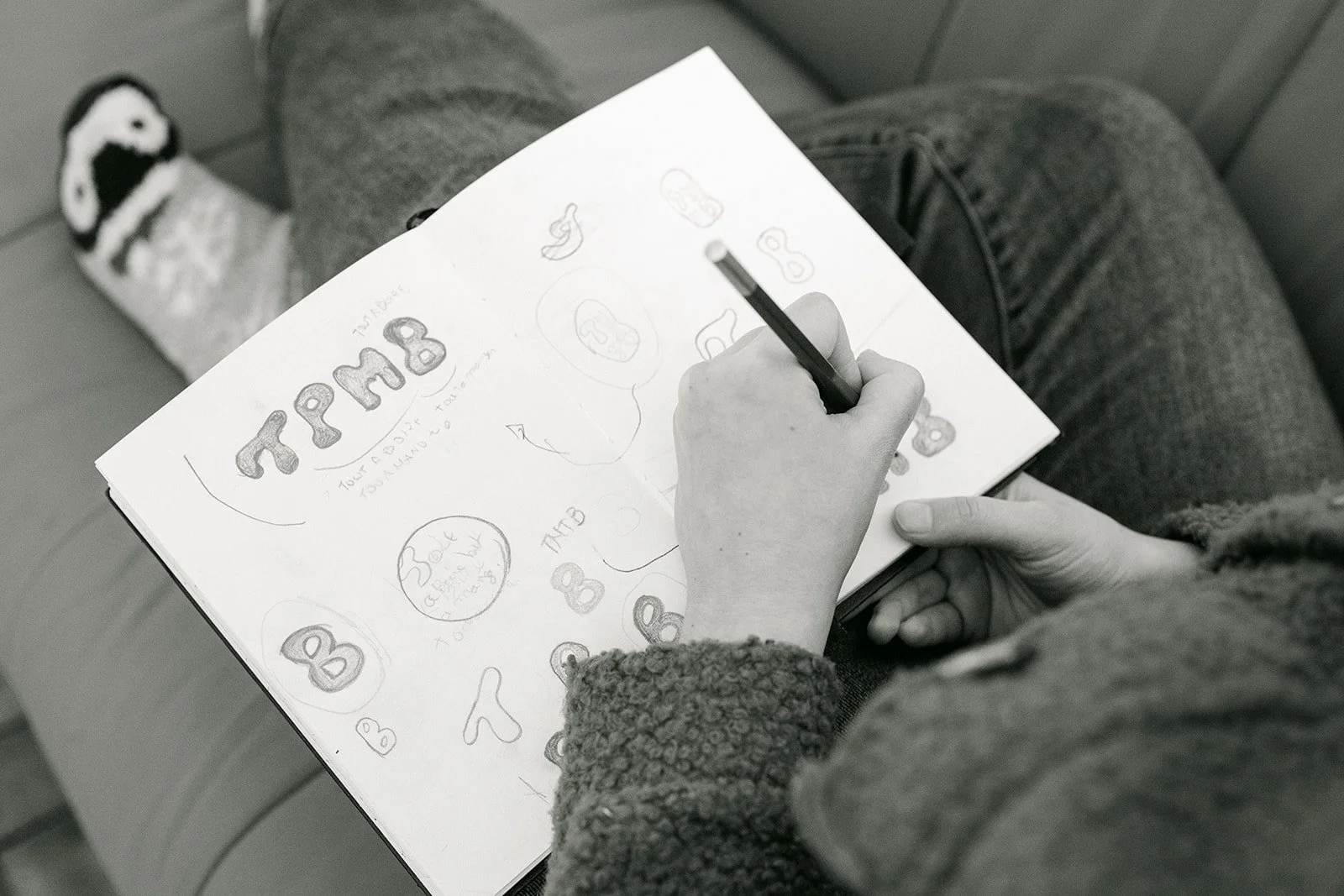 Une personne en train de faire des dessins et écrire sur un carnet. La page montre du texte "TTPMB" avec plusieurs dessins de lettres "B" autour, et une note manuscrite. La personne porte un pull en peluche et des jeans, et porte des chaussettes avec un dessin de chien.