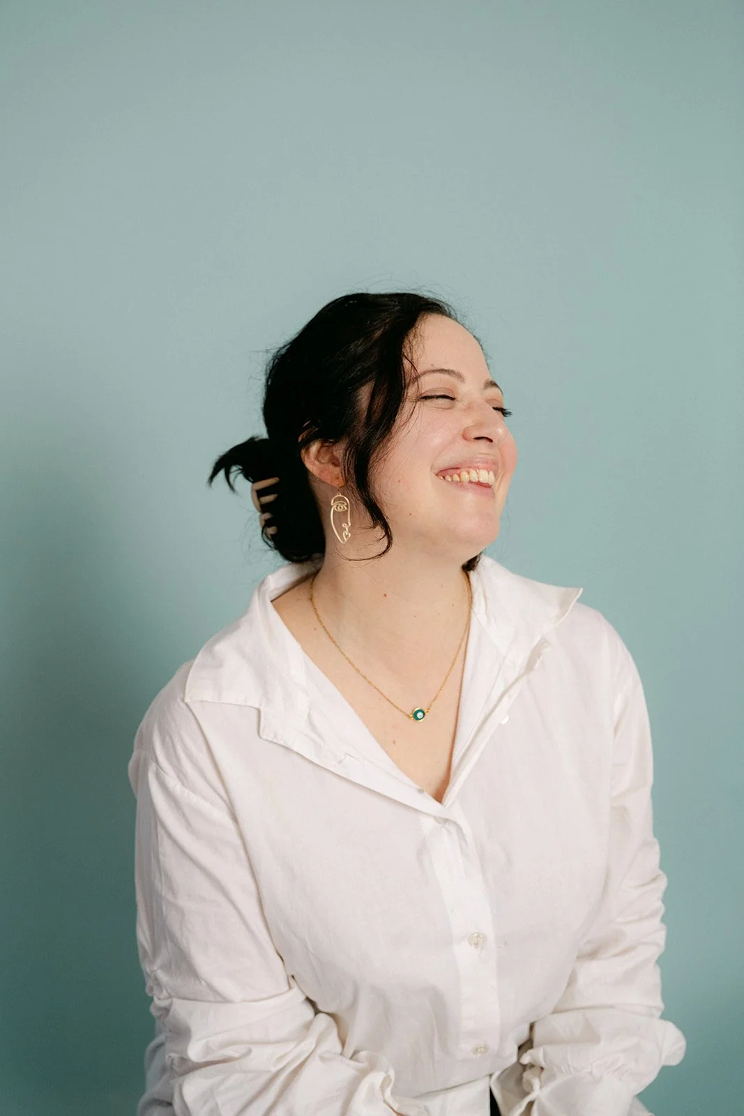 Femme souriante portant une chemise blanche et un jean, posant contre un mur blanc