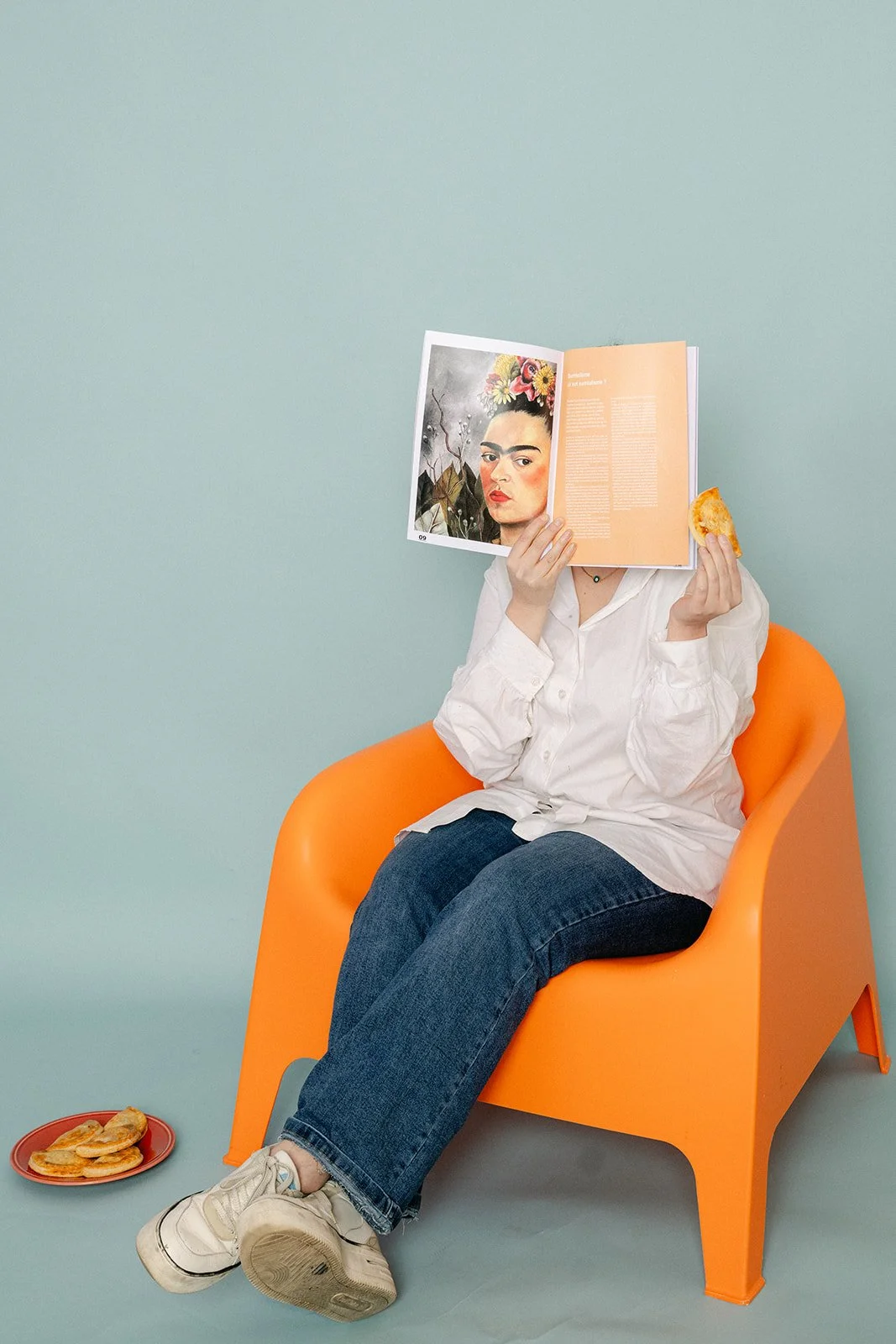 Une femme souriante, assise dans un fauteuil orange, portant une chemise blanche et des jeans, devant un mur bleu clair.
