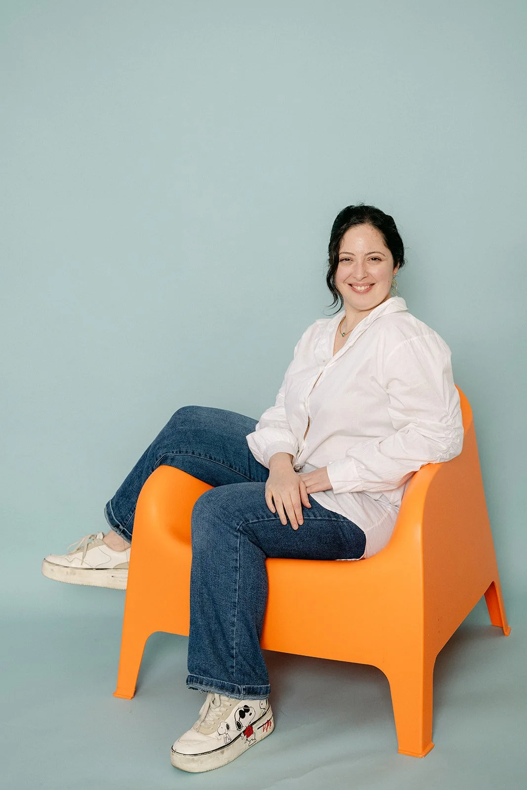 Une femme souriante, assise dans un fauteuil orange, portant une chemise blanche et des jeans, devant un mur bleu clair.