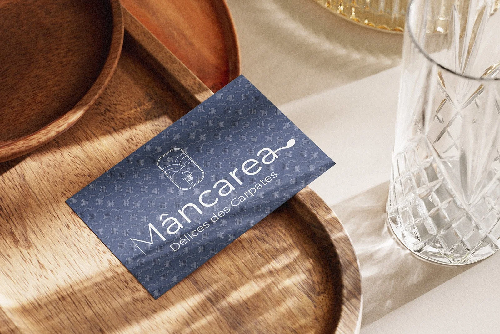 Une carte de visite bleue avec le texte 'Manara, Délices de Carpates' et une icône de mobylette, posée sur un plateau en bois à côté de deux verres en cristal.