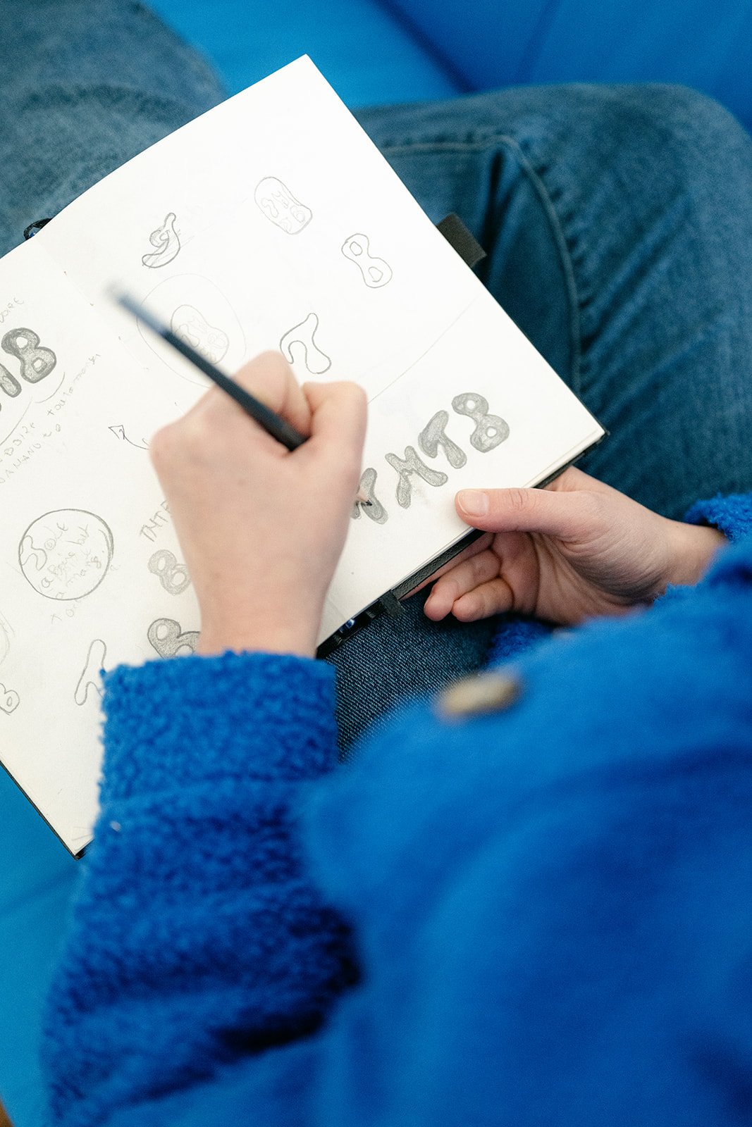 Une personne en pull bleu écrivant et dessinant différentes formes de chiffres 8 dans un carnet, assise avec des jeans bleus.