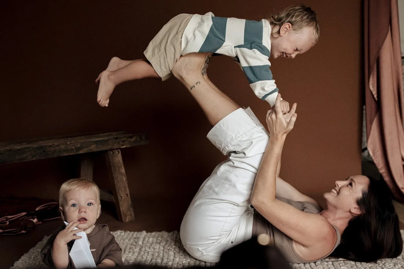 A woman with dark hair lying on her back on a rug, playing with a young boy on her legs, while a toddler looks on. The boy is airborne, being held by the woman, with a smile on the woman's face and the toddler holding a piece of paper.