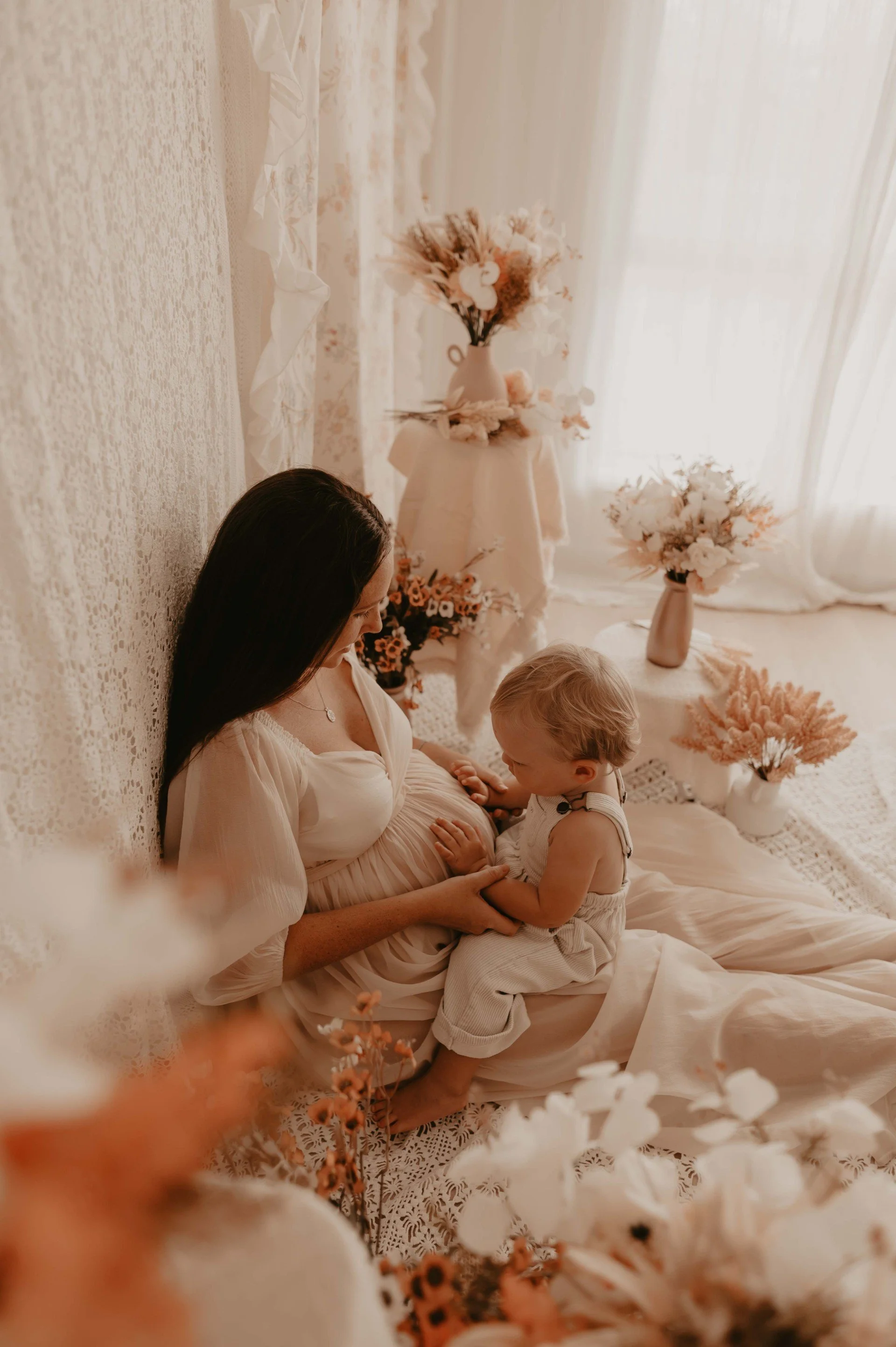 A woman with dark hair sitting on the floor in a cream dress, pregnant, holding a young child with blond hair, in a softly lit room with floral decorations and vases of flowers.