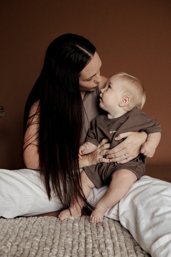 A woman and a young child sharing a close, affectionate moment indoors. The woman is sitting on a bed with a textured beige blanket, holding the child on her lap. They are looking at each other lovingly.