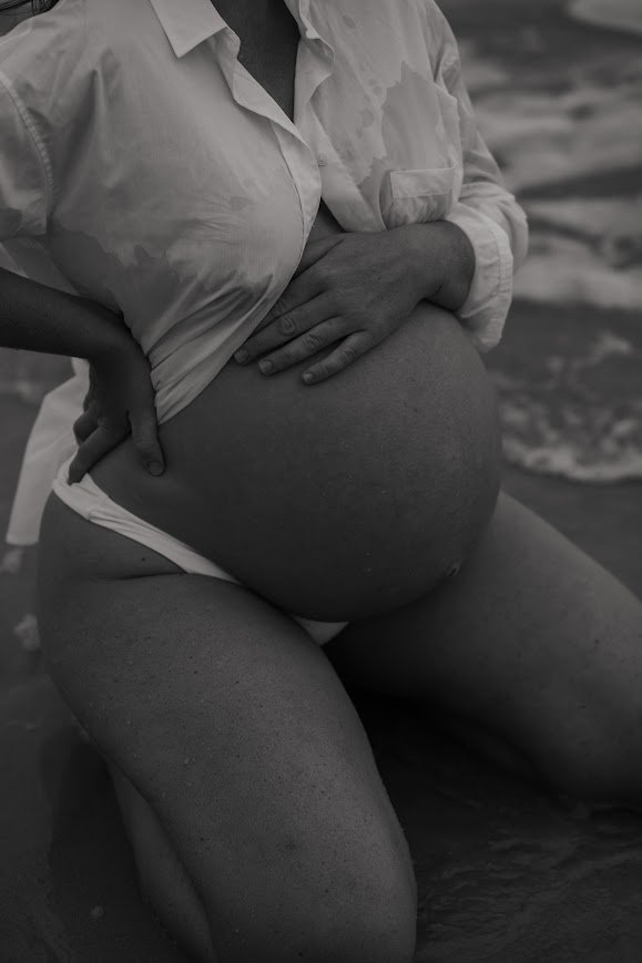 A pregnant woman in a white shirt and underwear kneeling on the beach with her hands on her belly.