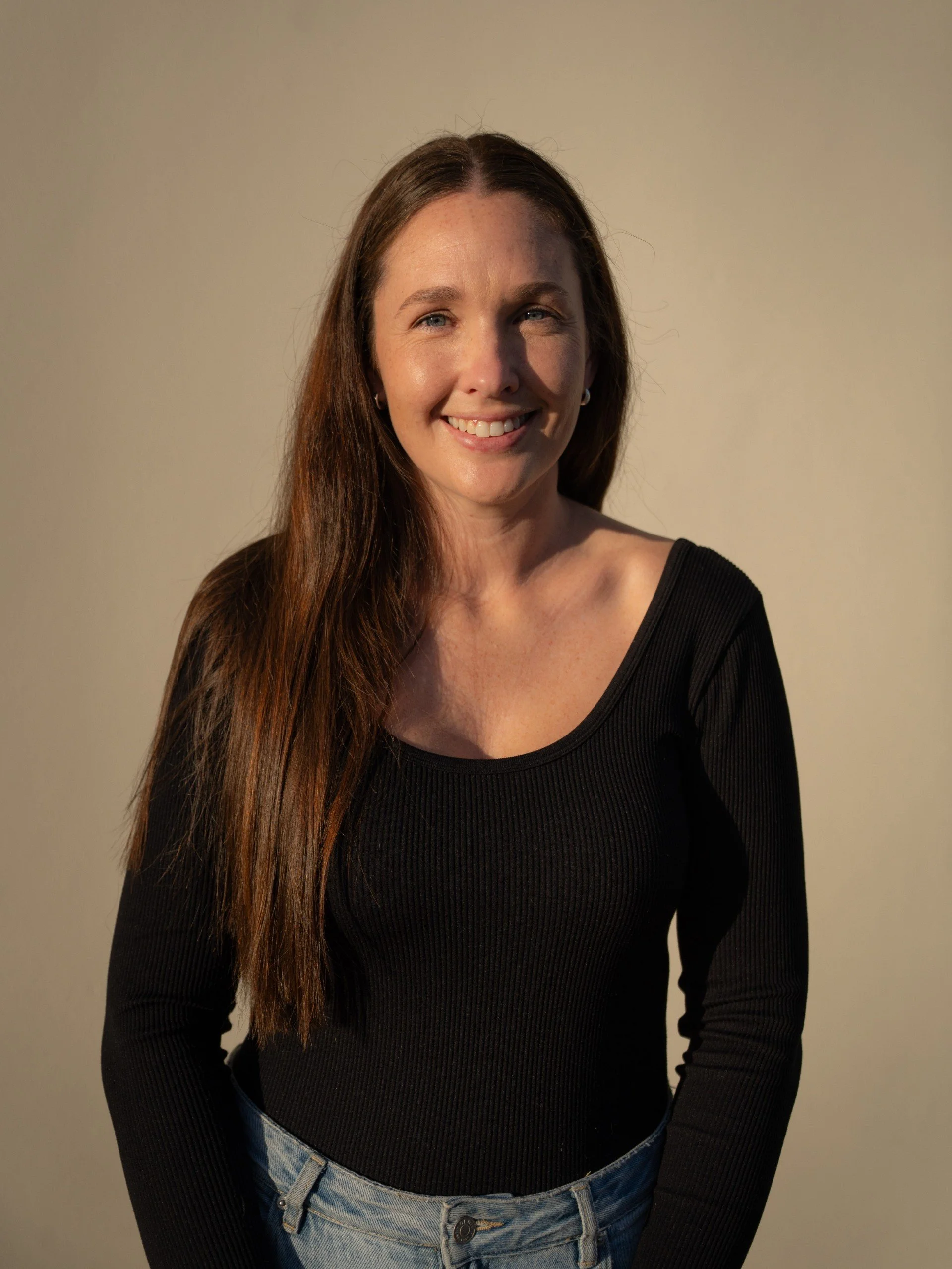 A woman with long brown hair, wearing a black long-sleeve shirt and blue jeans, smiling at the camera against a plain beige background.