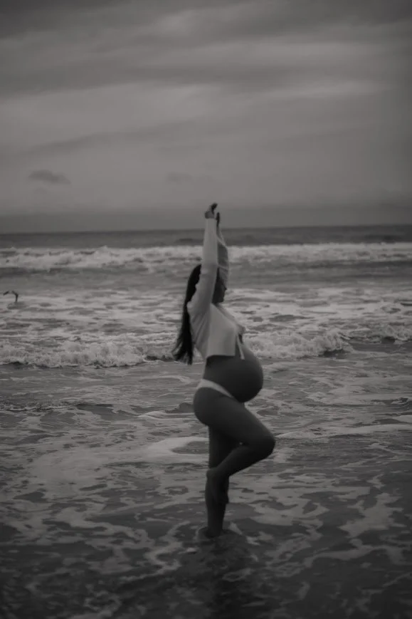 A pregnant woman practicing yoga on the beach, standing on one leg with arms raised above her head, holding an object, with the ocean and cloudy sky in the background.