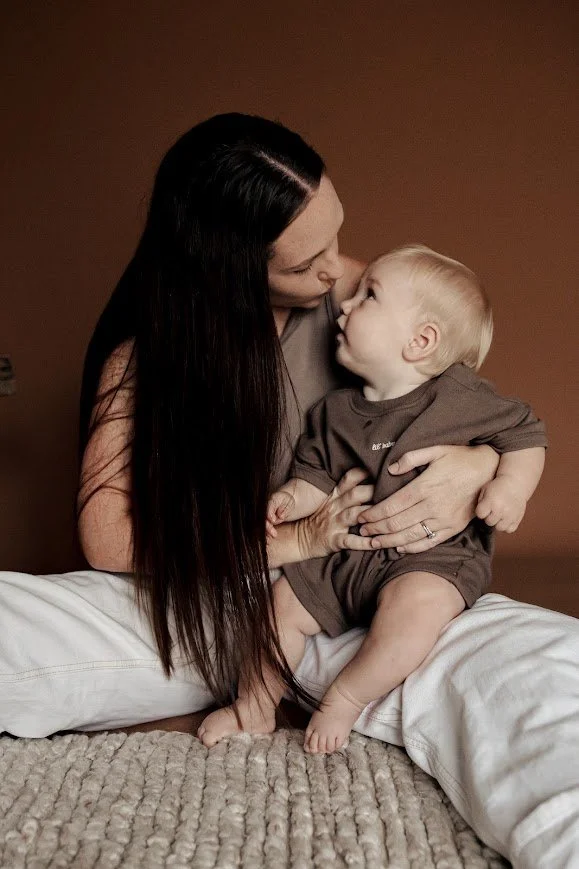 A woman with long dark hair holding a young blonde boy, both sitting on a carpeted floor against a brown background, sharing a close, affectionate moment.