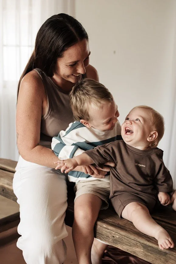 Woman sitting on a wooden surface with two young boys, one boy is leaning toward her, the other boy is sitting and laughing, all are enjoying a joyful moment together.